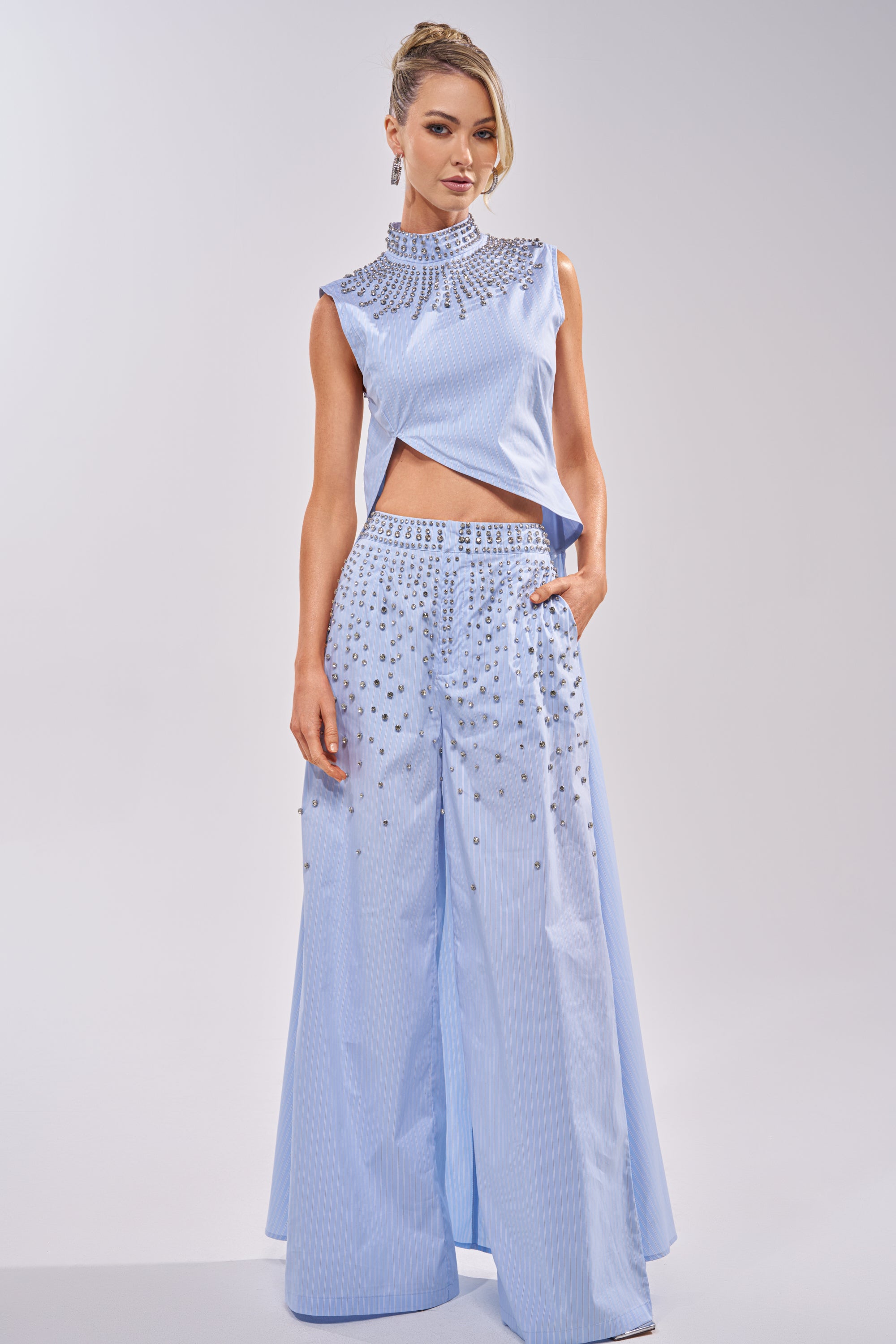 A woman poses in the STAR OF THE NIGHT POPLIN BLOUSE, a sleeveless light blue crop top with a rhinestone collar and intricate beading, paired with matching wide-leg pants. Her hair is styled up against a plain, light background. 0