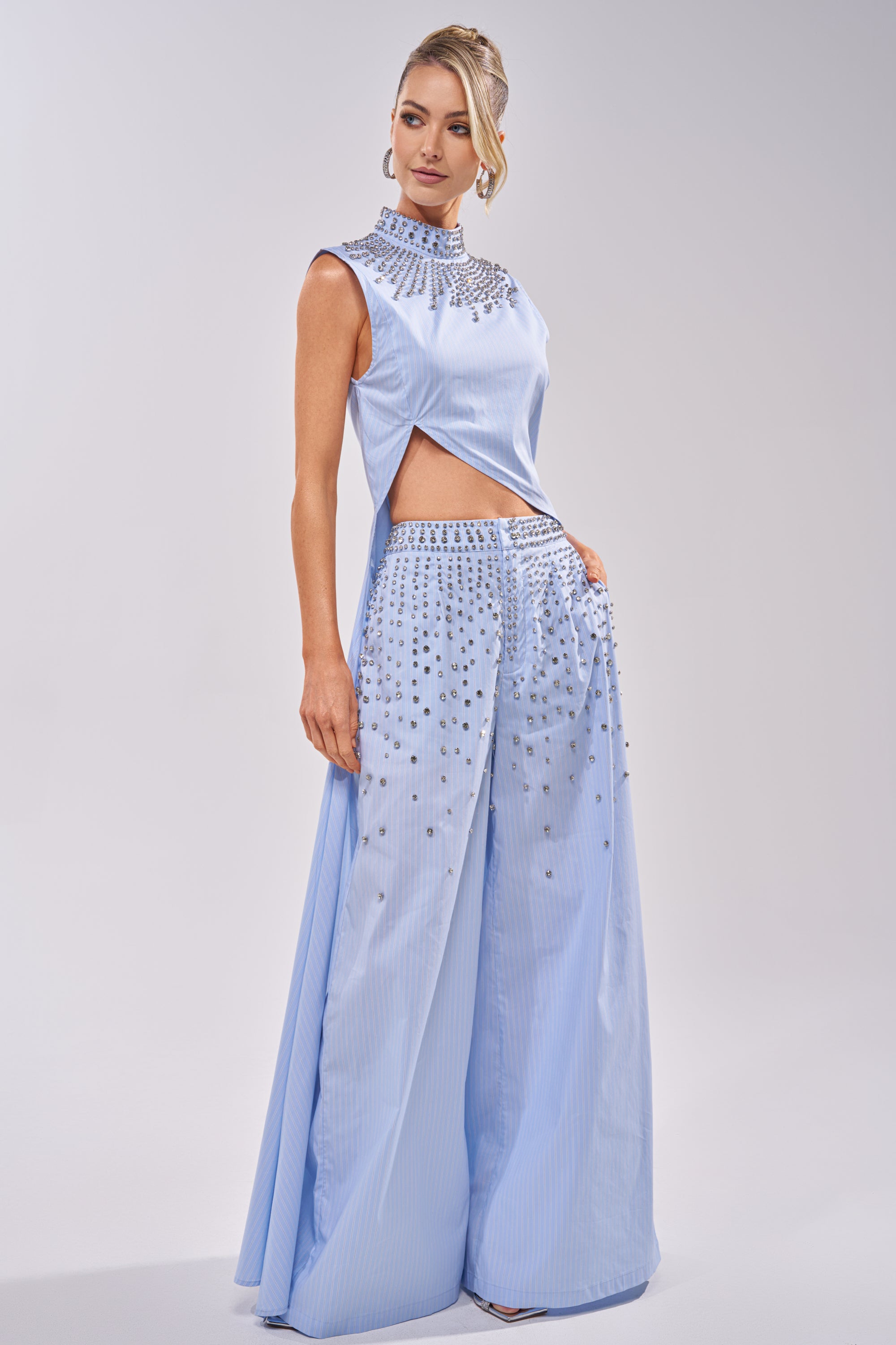 A woman stands against a plain background in the STAR OF THE NIGHT POPLIN BLOUSE—light blue, sleeveless, with a rhinestone collar and side cutouts—paired with wide-leg pants featuring silver embellishments and an elegant updo. 1