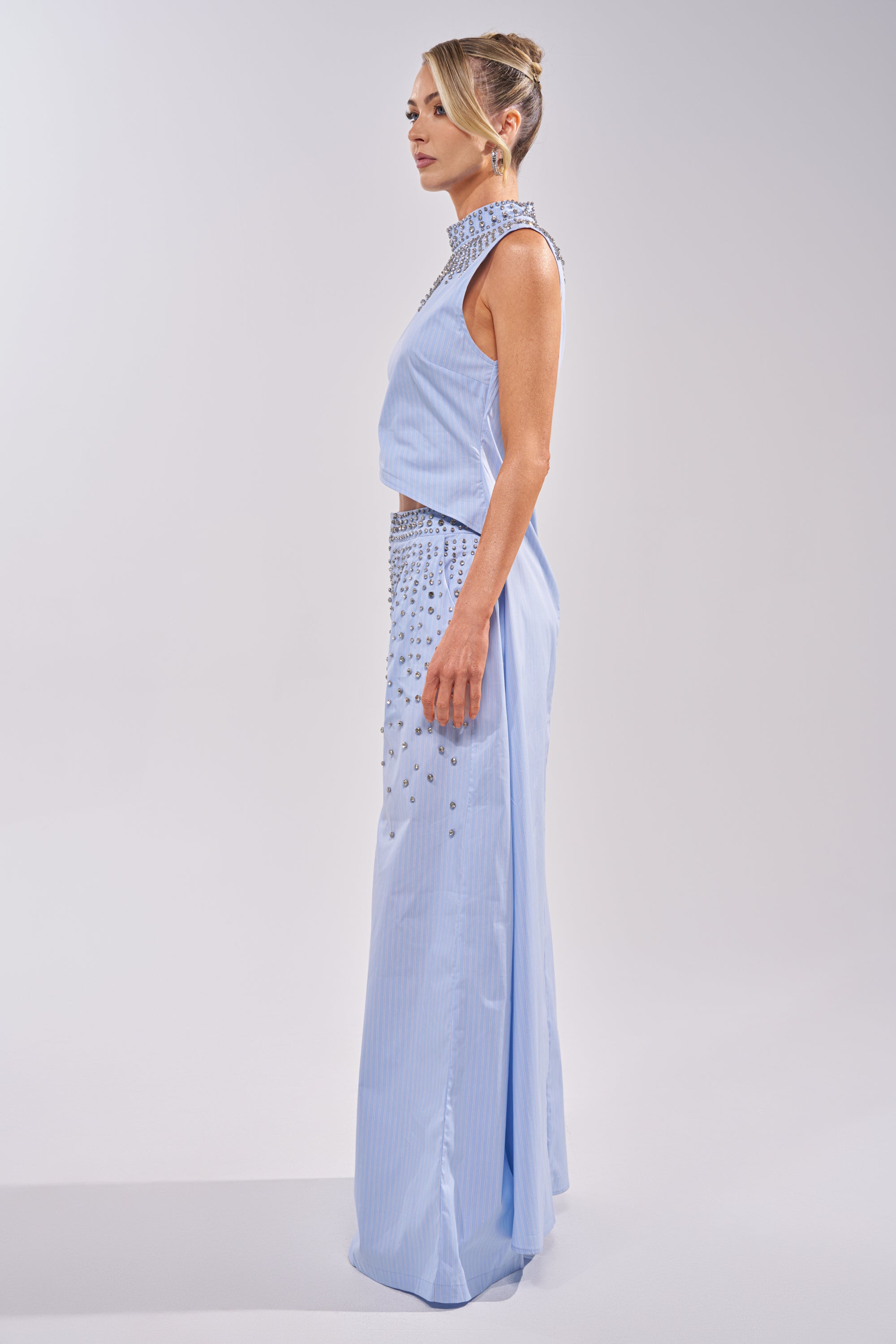 A woman in profile wears the STAR OF THE NIGHT POPLIN BLOUSE, a sleeveless, light blue top with a rhinestone collar and silver accents at the waist. Her hair is in an elegant updo against a plain, light-colored background. 2