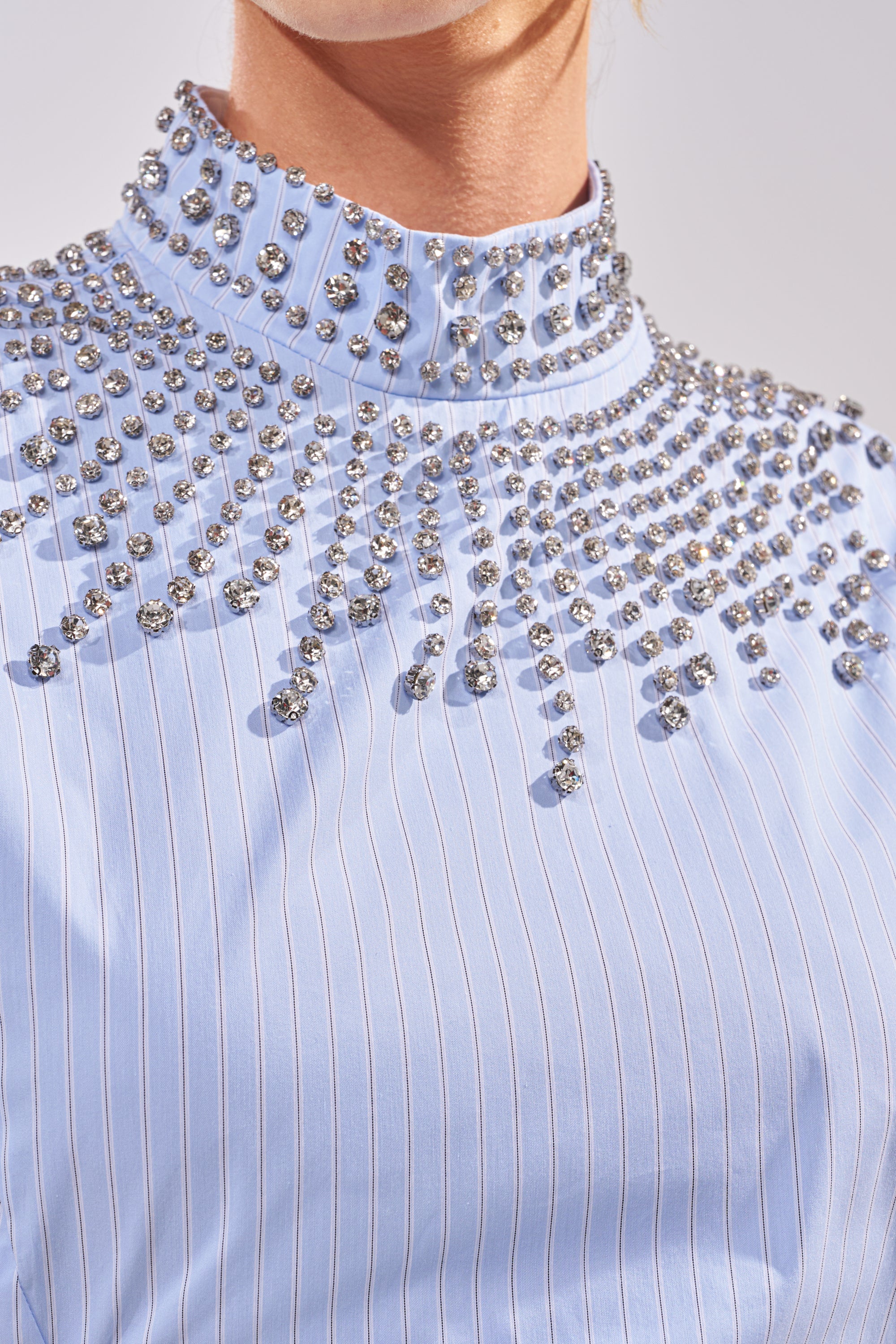 Close-up of the STAR OF THE NIGHT POPLIN BLOUSE, a light blue pinstripe blouse with a high collar and rows of large, round rhinestones radiating from the collar and shoulders, worn by a person. 5