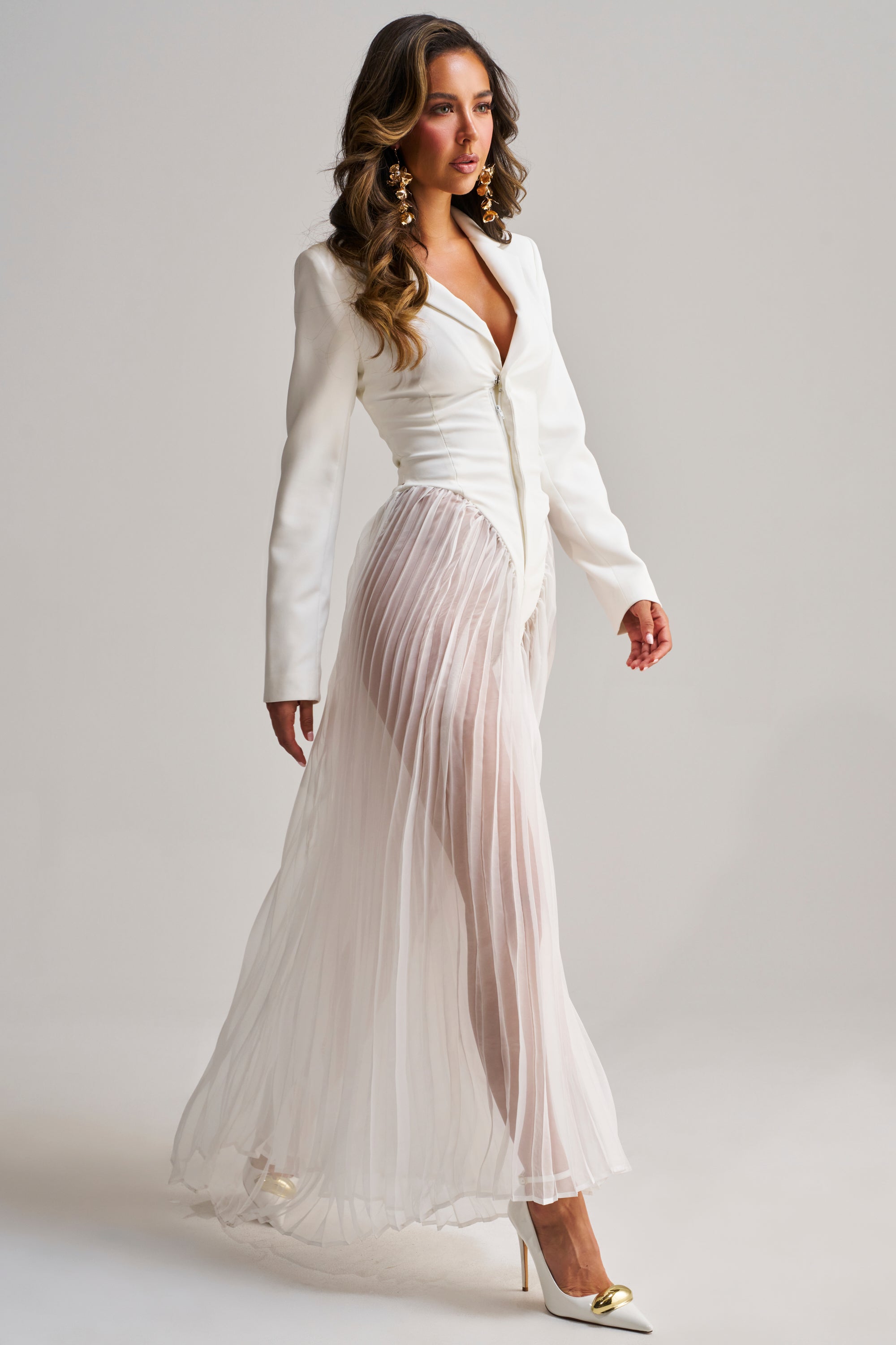 A woman with long wavy hair walks confidently in the ICON BEHAVIOR PLEATED BLAZER JUMPSUIT, featuring a sheer, pleated chiffon skirt and styled with white high heels with gold accents, set against a plain background.