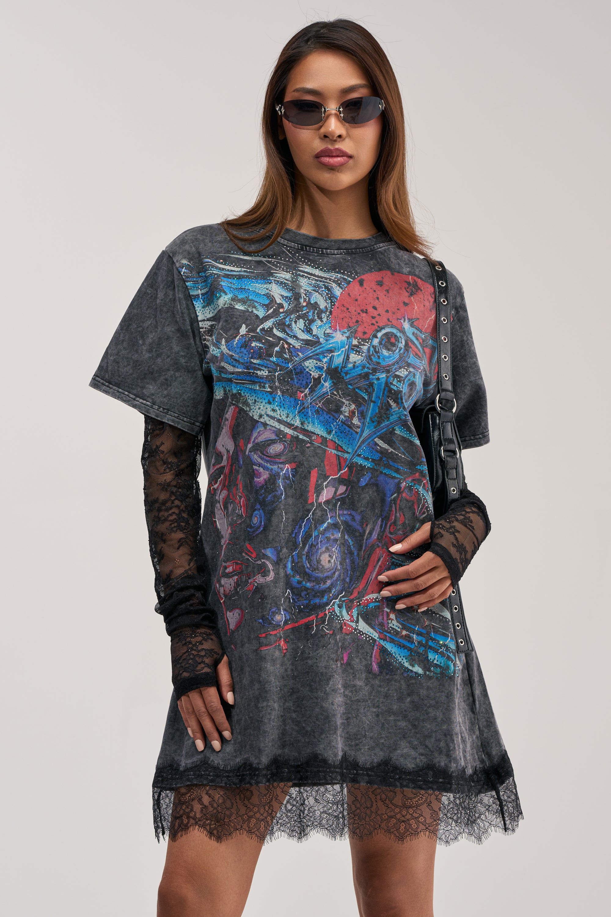 Model is wearing the THEY WATCH I WIN GRAPHIC LACE MINI DRESS—a rhinestone-embellished T-shirt mini dress featuring long lace sleeves and lace trim—styled with sunglasses and a black purse against a light backdrop. This eye-catching piece makes the perfect concert outfit or standout streetwear look.