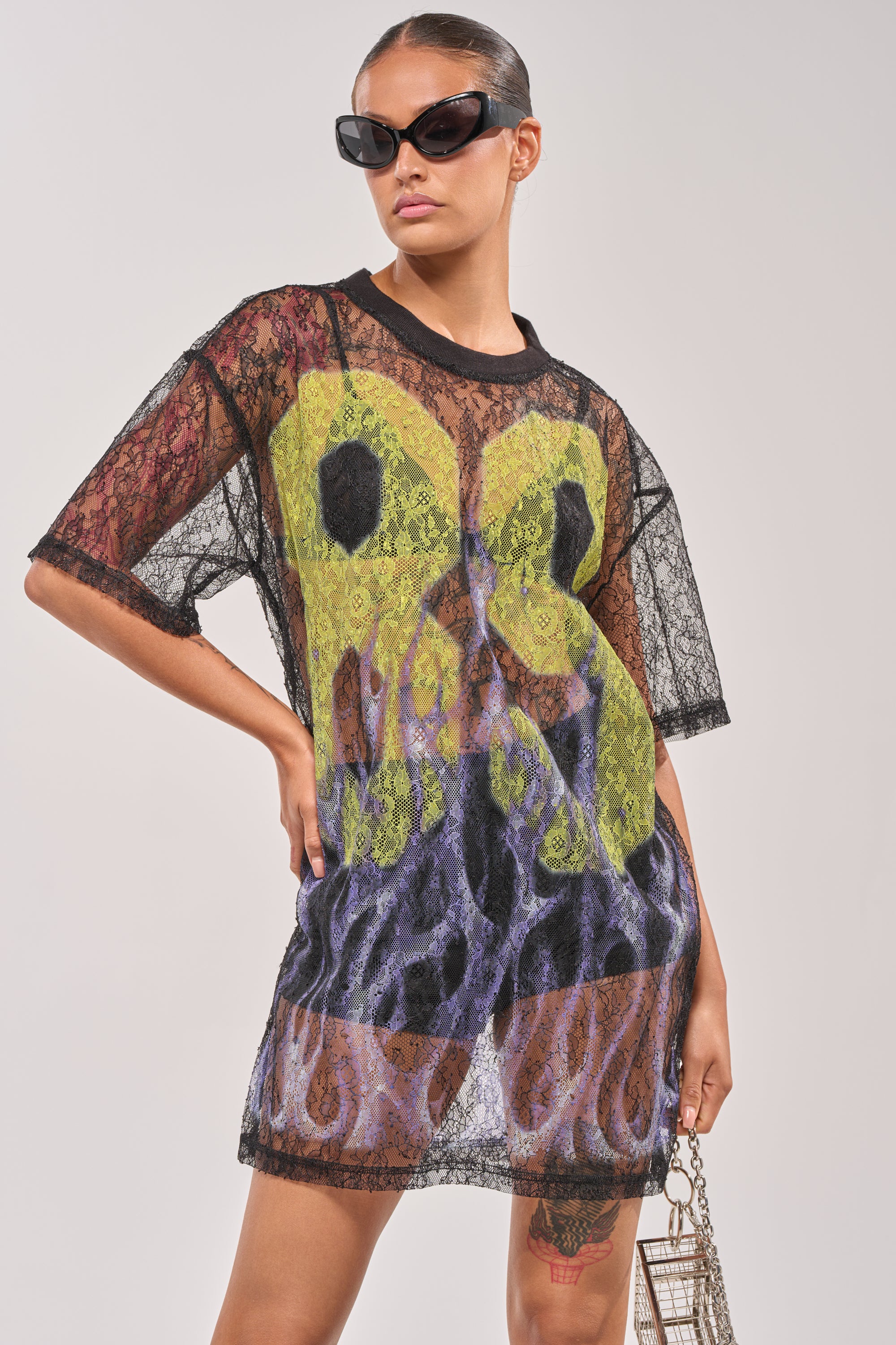 A confident person wears the ALL THE RAGE LACE JERSEY MINI DRESS—sheer, short-sleeved, and black with striking yellow and purple flame designs—paired with black sunglasses and a small metallic purse. 1