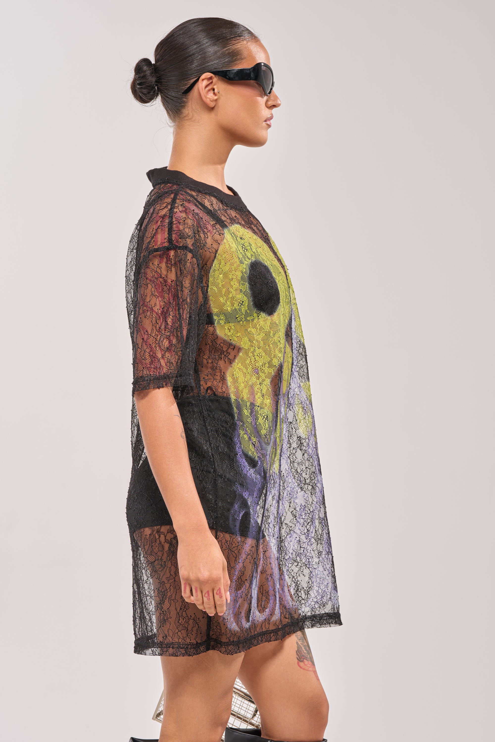 A woman with her hair in a bun and sunglasses poses in profile, wearing the ALL THE RAGE LACE JERSEY MINI DRESS over a graphic top and black shorts against a plain light background. 2