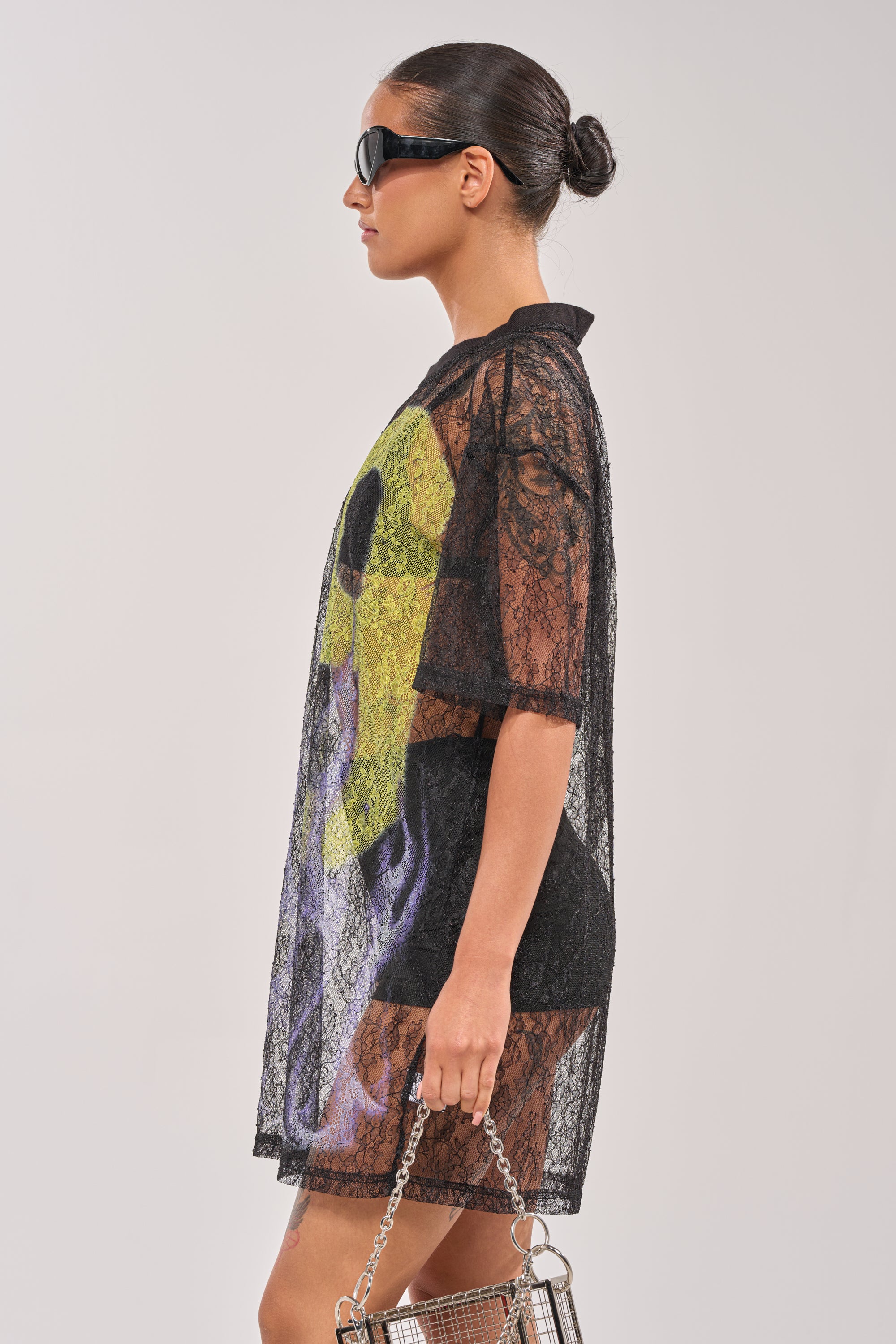 A woman in profile wears the ALL THE RAGE LACE JERSEY MINI DRESS over a colorful outfit, paired with dark sunglasses and a small metallic handbag. Her hair is styled in a sleek bun against a plain light background. 3