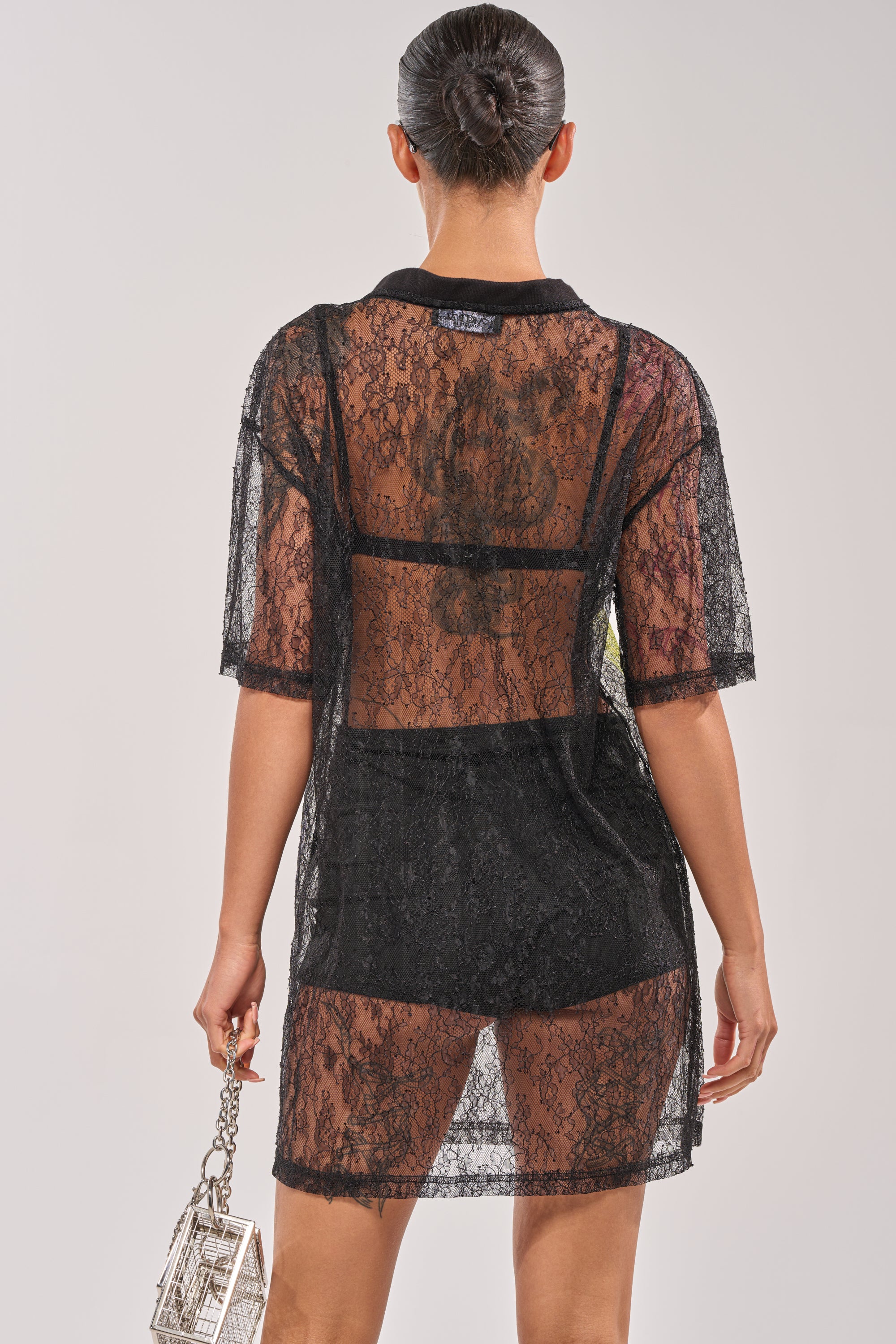 A person with hair in a neat bun is shown from the back, wearing the ALL THE RAGE LACE JERSEY MINI DRESS over black undergarments and holding a small, boxy handbag with a metallic handle. 4