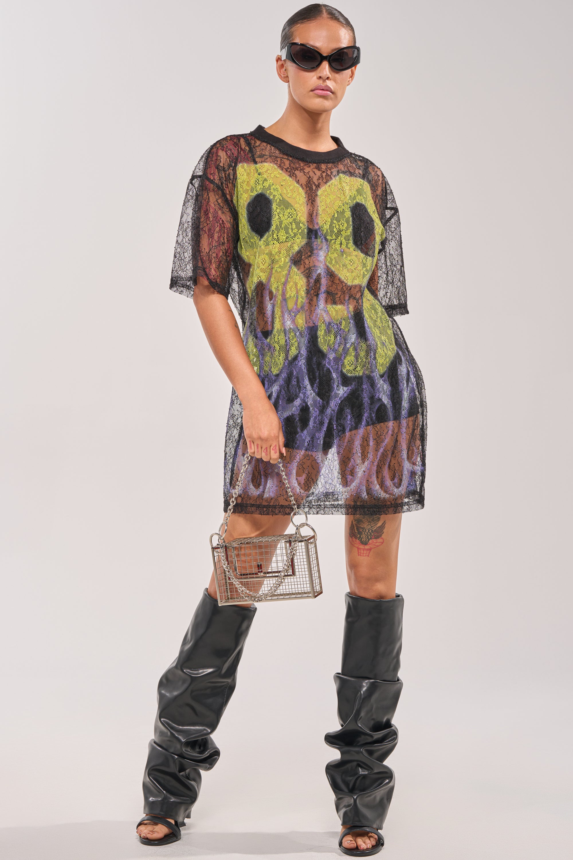 Wearing sunglasses, a person models the ALL THE RAGE LACE JERSEY MINI DRESS—sheer black mesh with green and purple abstract patterns—over a slip, paired with black slouchy knee-high boots and a small box handbag against a plain background. 5