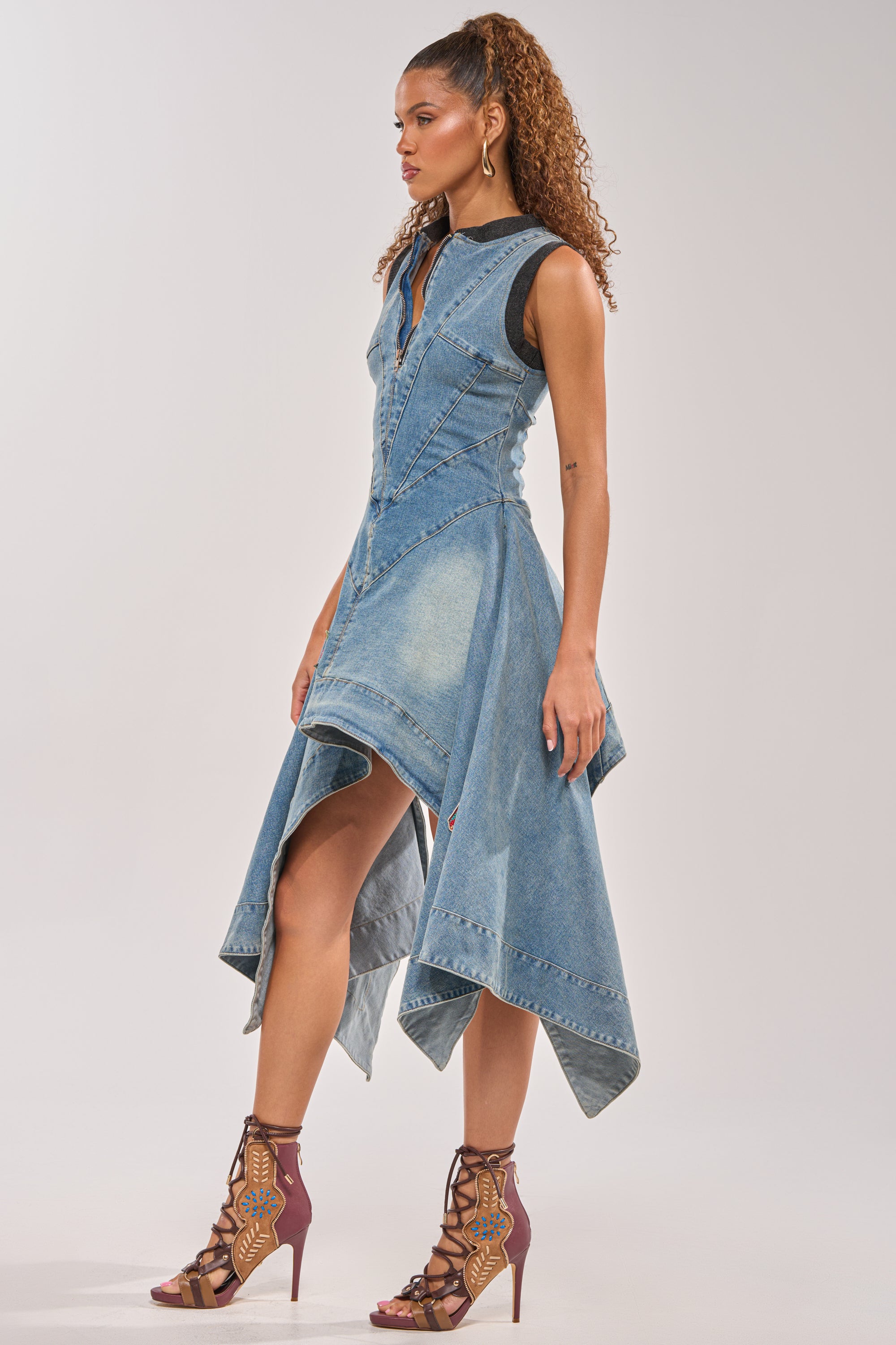 A woman models the BALLER OFF DUTY DENIM PATCHWORK MINI DRESS, a sleeveless mini with an asymmetrical hem and front zipper, styled with colorful lace-up high-heeled sandals featuring playful embroidered patches. 1