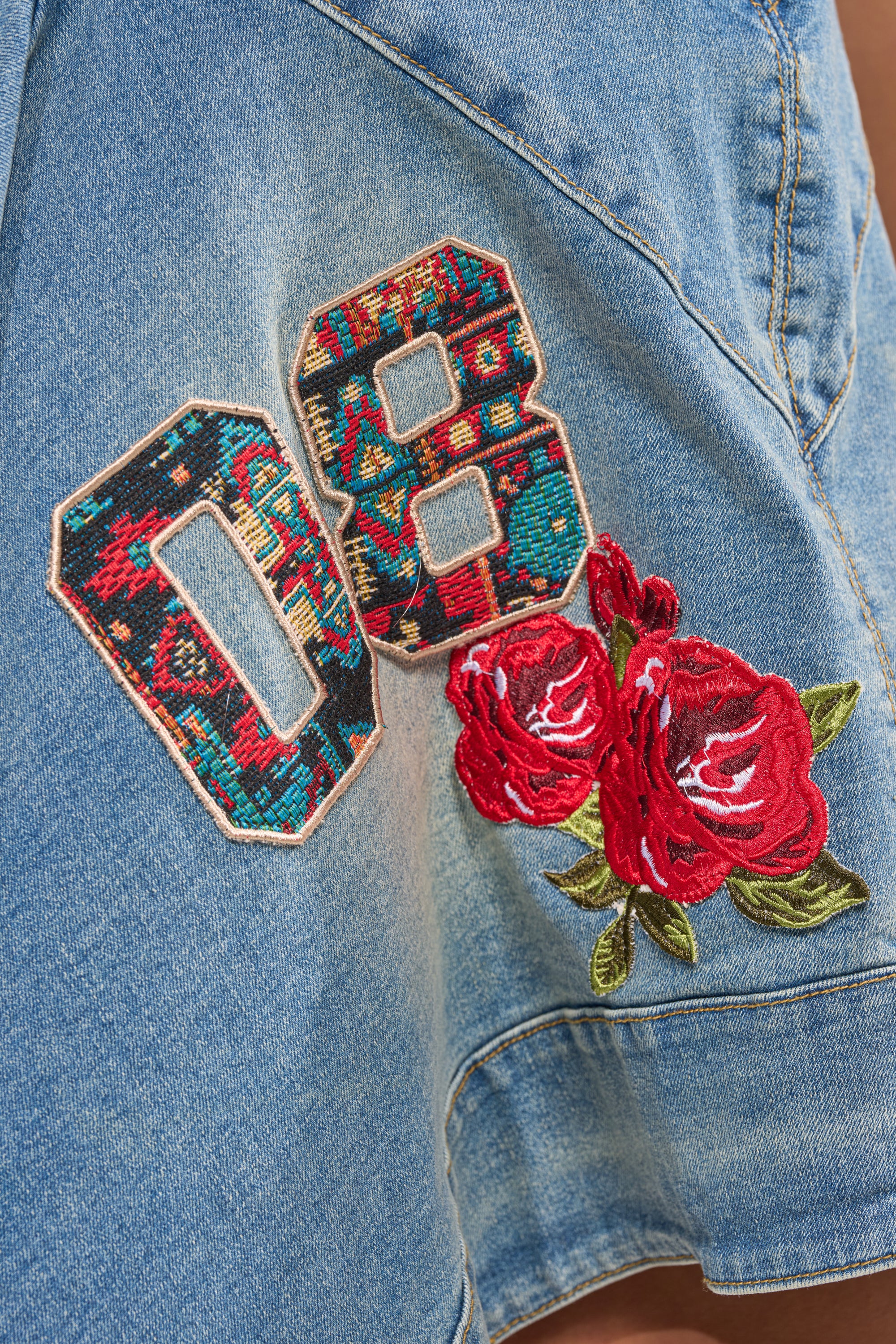 Close-up of the BALLER OFF DUTY DENIM PATCHWORK MINI DRESS in light blue denim, featuring embroidered "08" patches in vibrant colors and two red rose appliqués with green leaves. 4