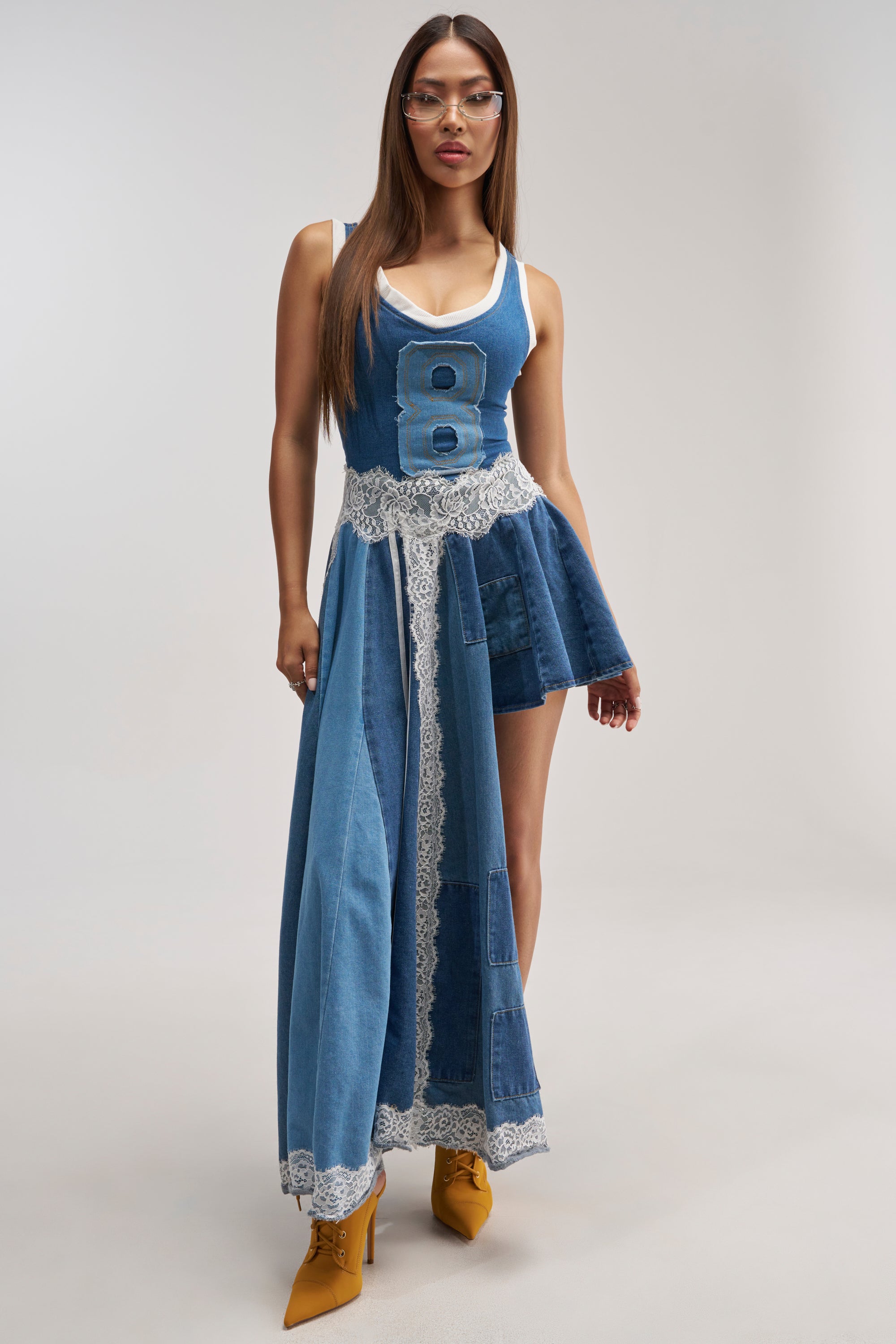 A woman wearing the BOSSED UP DENIM MINI DRESS IN LIGHT BLUE DENIM PATCHWORK—an asymmetrical denim patchwork mini dress with lace trim and a number 8 on the bodice—pairs it with yellow heels and glasses as she stands against a plain background.