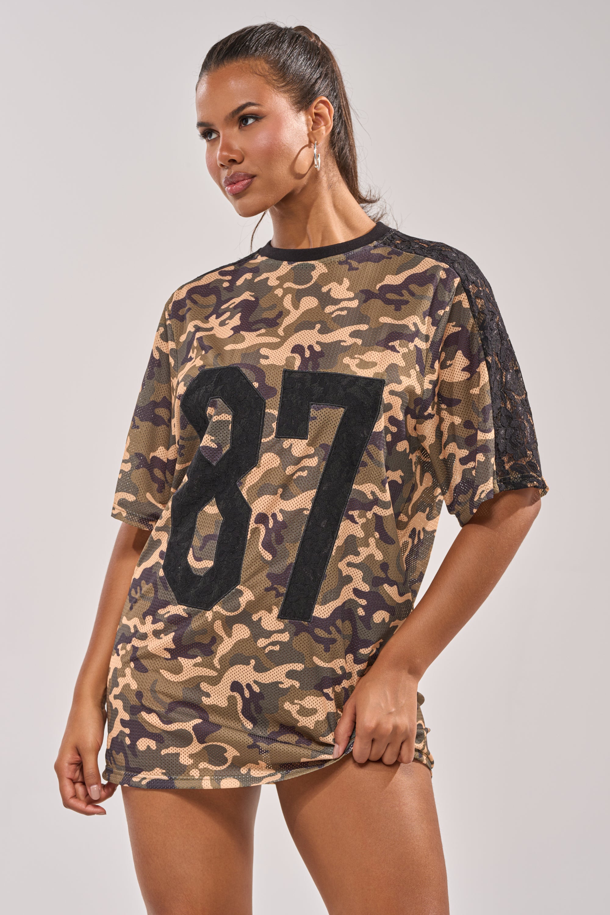 A woman poses against a plain background in the POPPIN POPPYS Lace Jersey Mini Dress in camo, featuring a sporty-chic loose fit, black lace on one sleeve, and a bold black "87" on the front. 2