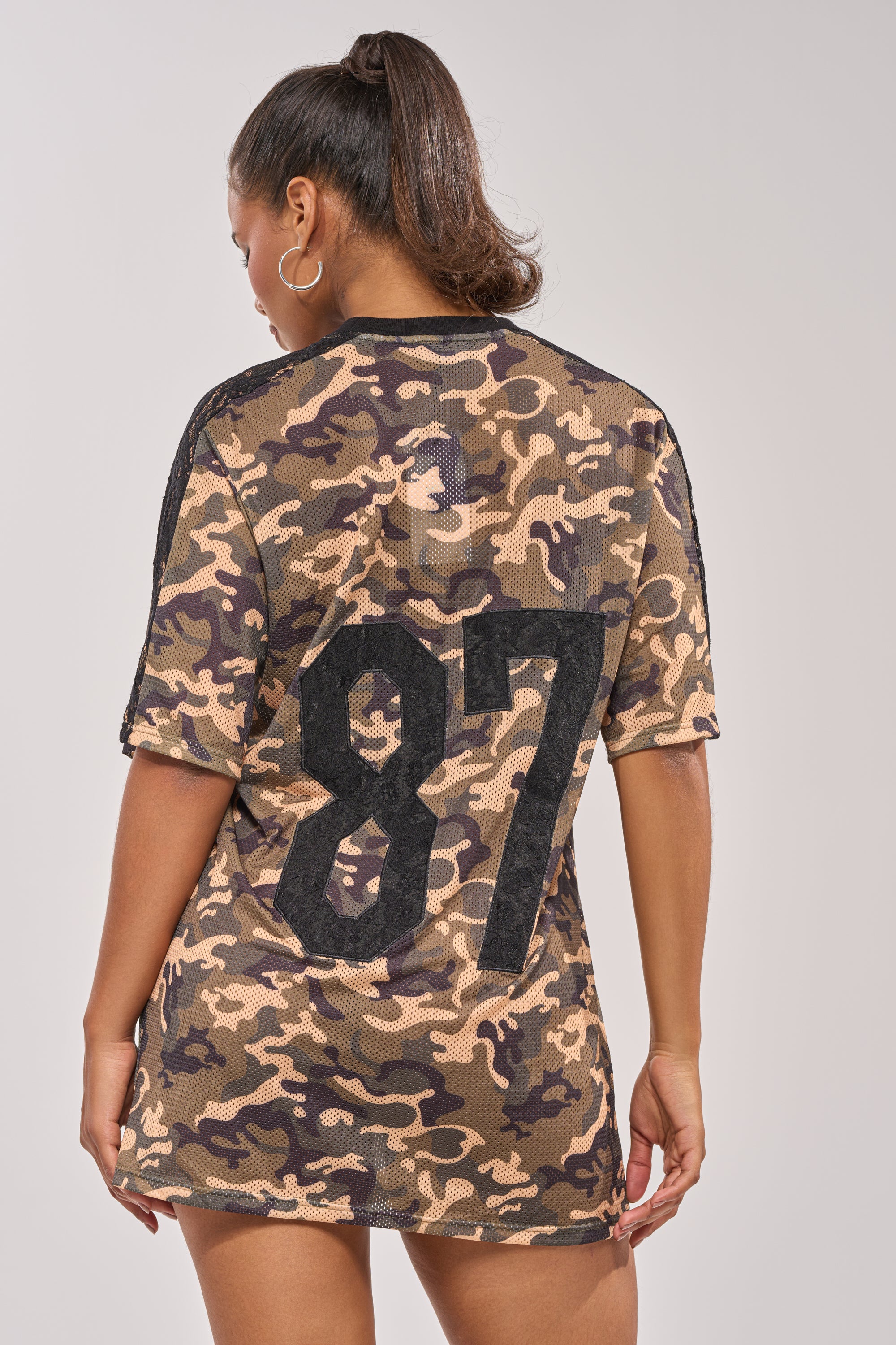 A woman faces away in the POPPIN POPPYS LACE JERSEY MINI DRESS IN CAMO, featuring sporty mesh fabric and a bold "87" on the back. Her ponytail and hoop earrings complete the cool athletic look. 5