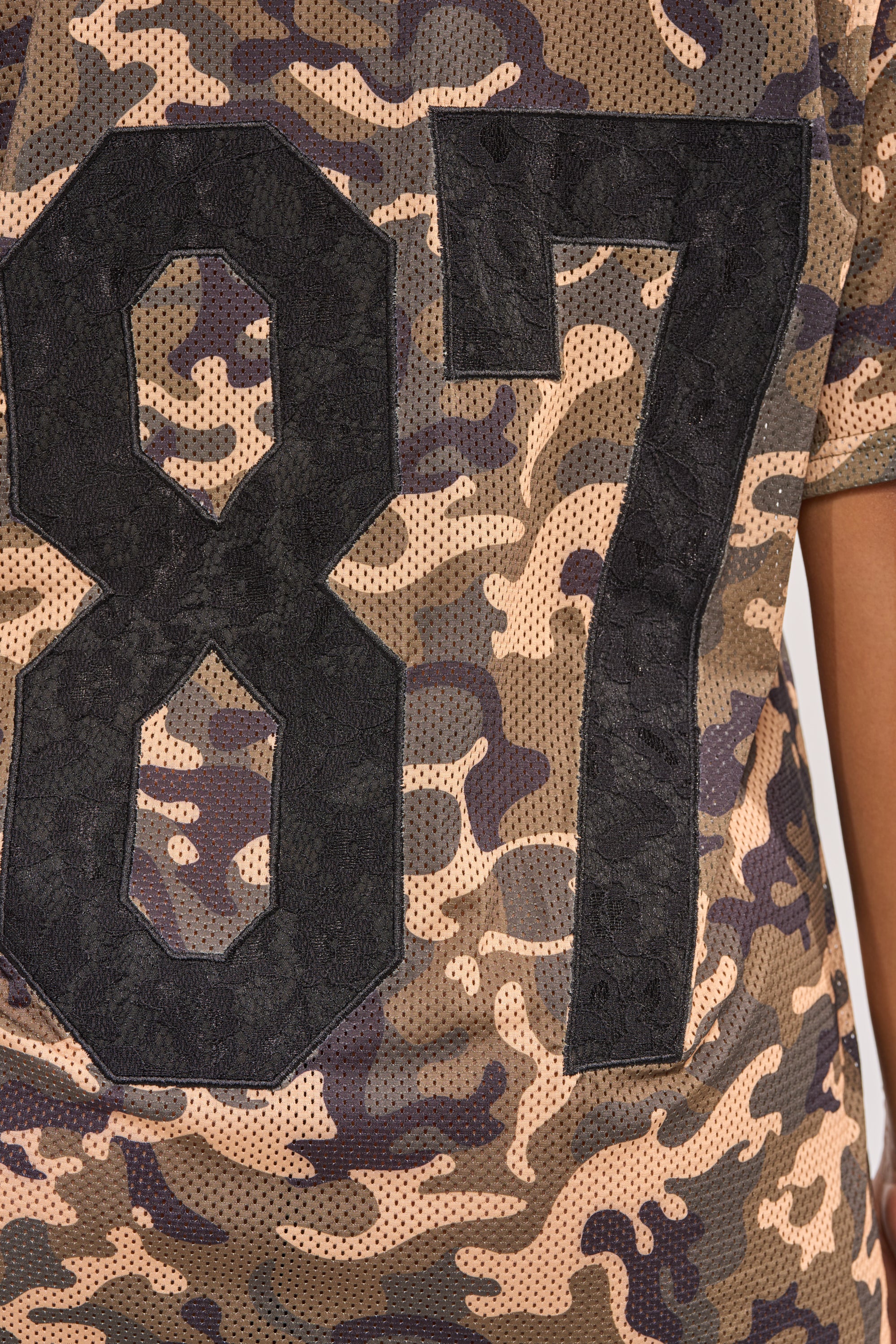 A close-up of the POPPIN POPPYS LACE JERSEY MINI DRESS IN CAMO, showcasing large black “87” numbers on the back. The sporty-chic camo dress is modeled, with only the person’s right arm partially showing. 8