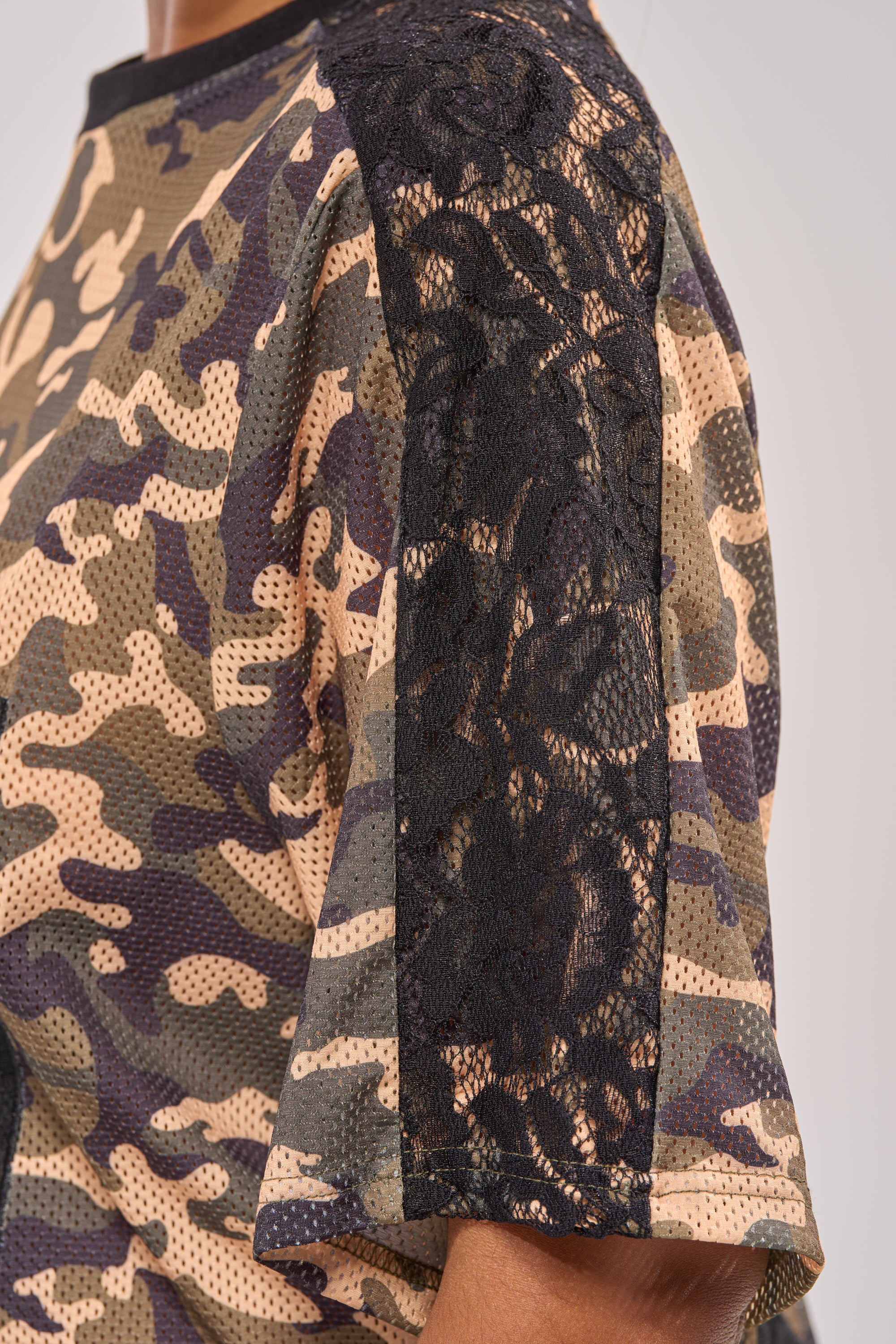 A close-up of a person wearing the POPPIN POPPYS LACE JERSEY MINI DRESS IN CAMO, featuring black lace on the shoulder and sleeve. The focus is on the upper torso and arm against a neutral background. 9