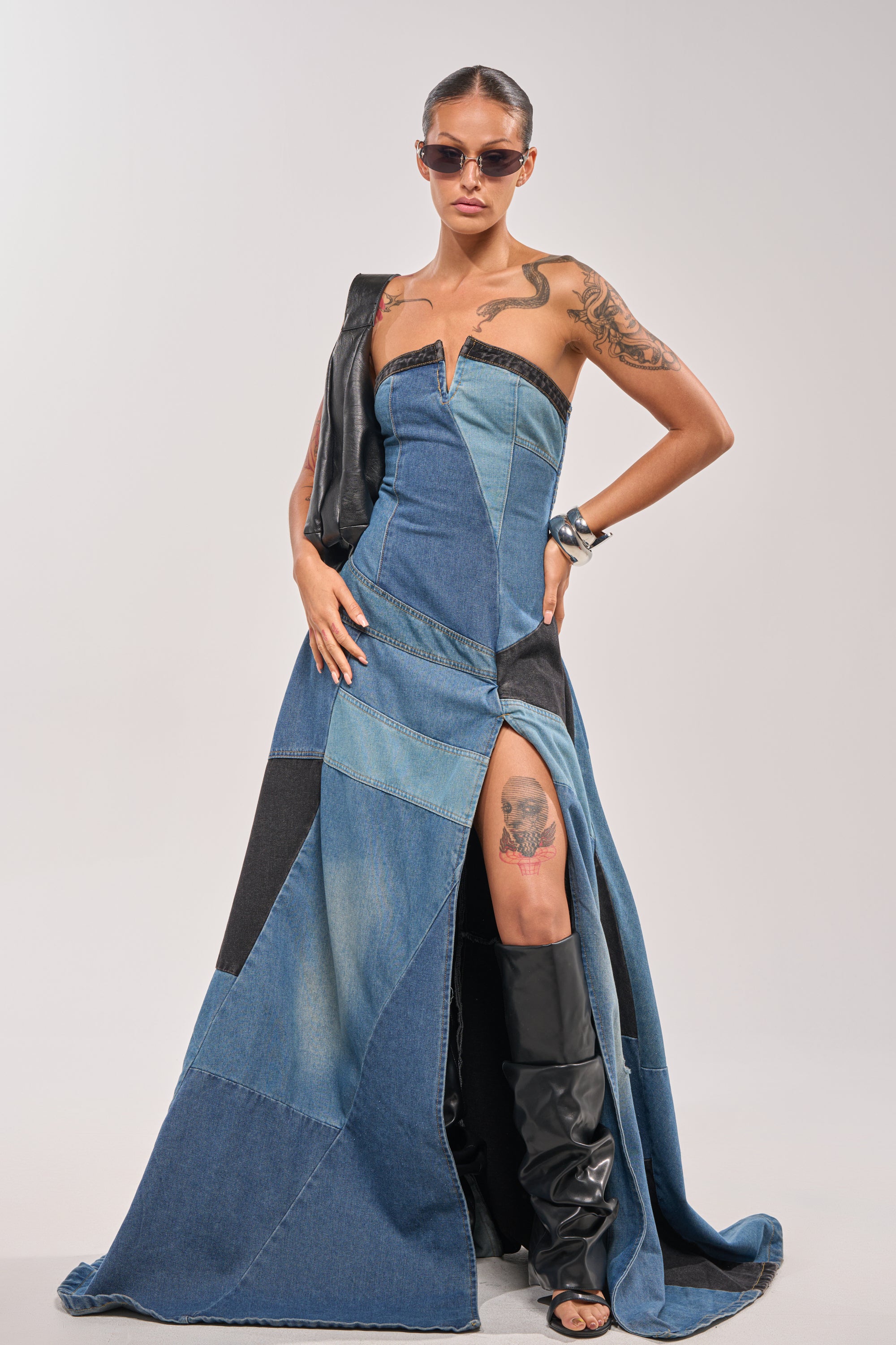 A person poses confidently in the LIFE IN DENIM STRAPLESS MAXI DRESS, featuring patchwork details and a high slit. Styled with black heeled boots, sunglasses, and sleek hair, this look is completed by a black leather accessory over one shoulder. 0