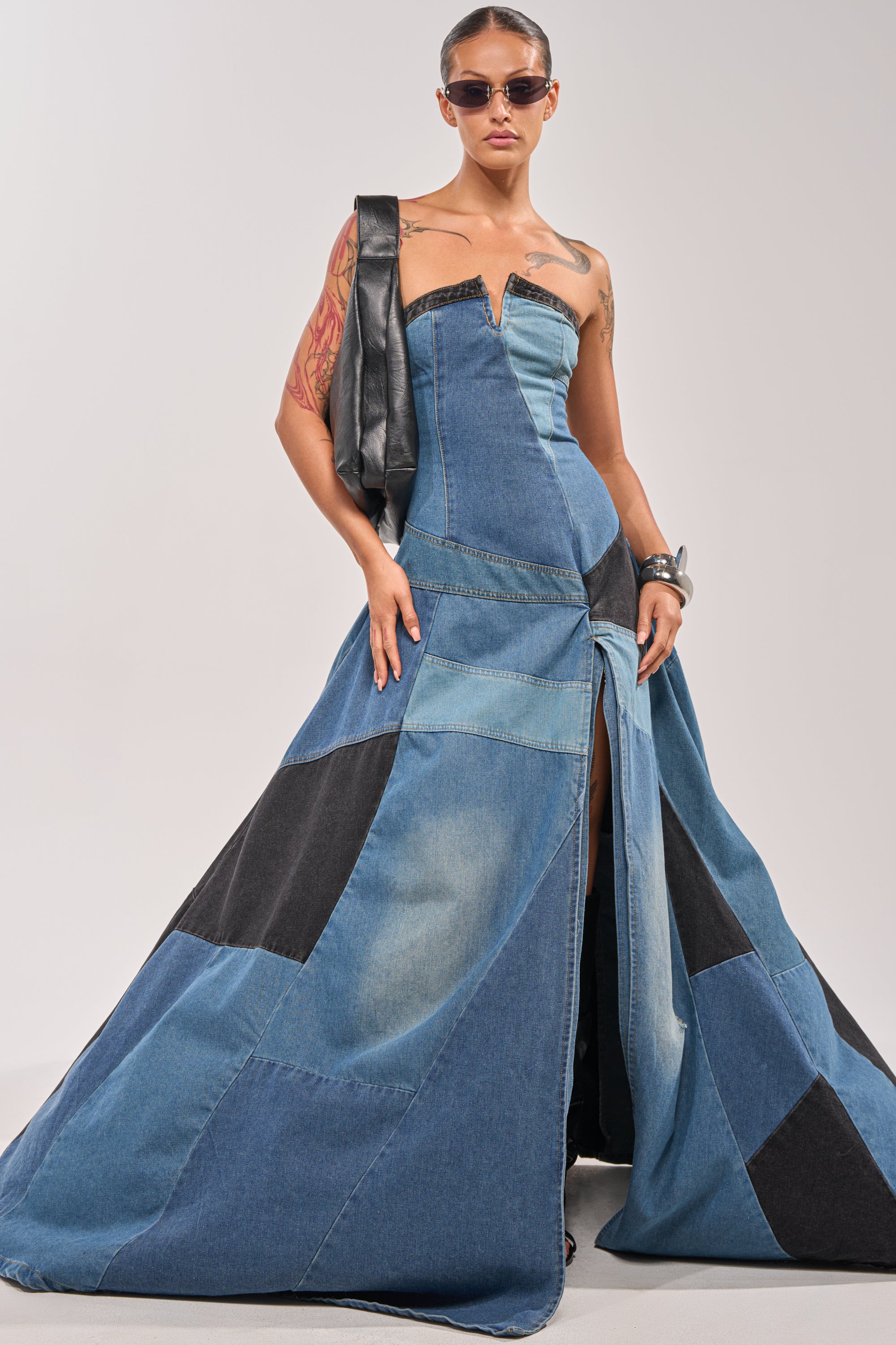 A model wears the LIFE IN DENIM STRAPLESS MAXI DRESS with patchwork details and a high slit, paired with sunglasses, chunky bracelets, and a black sculptural bag—modern evening wear reimagined against a plain backdrop. 1