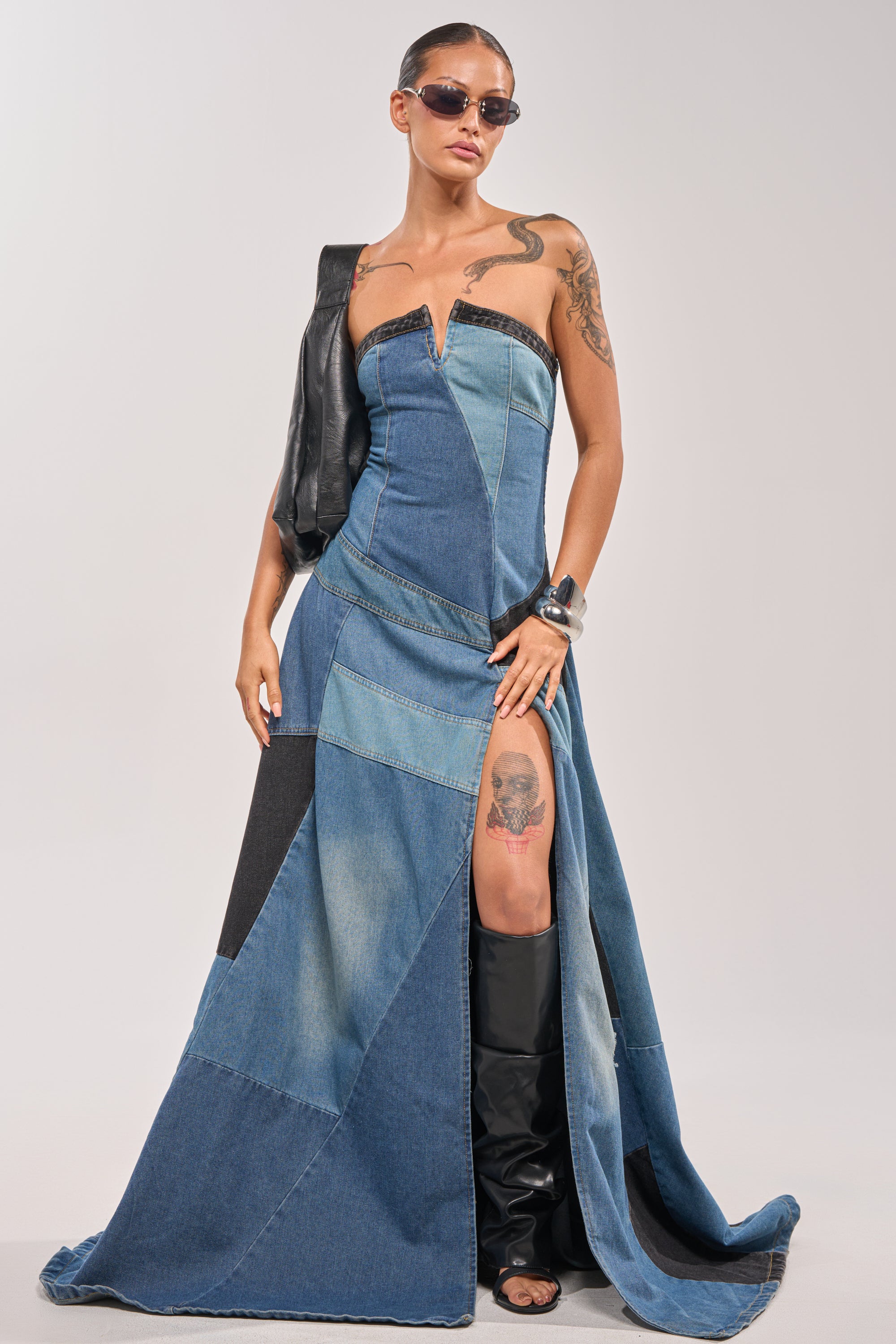 A woman stands confidently in the LIFE IN DENIM STRAPLESS MAXI DRESS, featuring a thigh-high slit. She pairs it with sunglasses, black boots, and a leather jacket over one shoulder—showcasing arm and thigh tattoos for an edgy evening look. 2