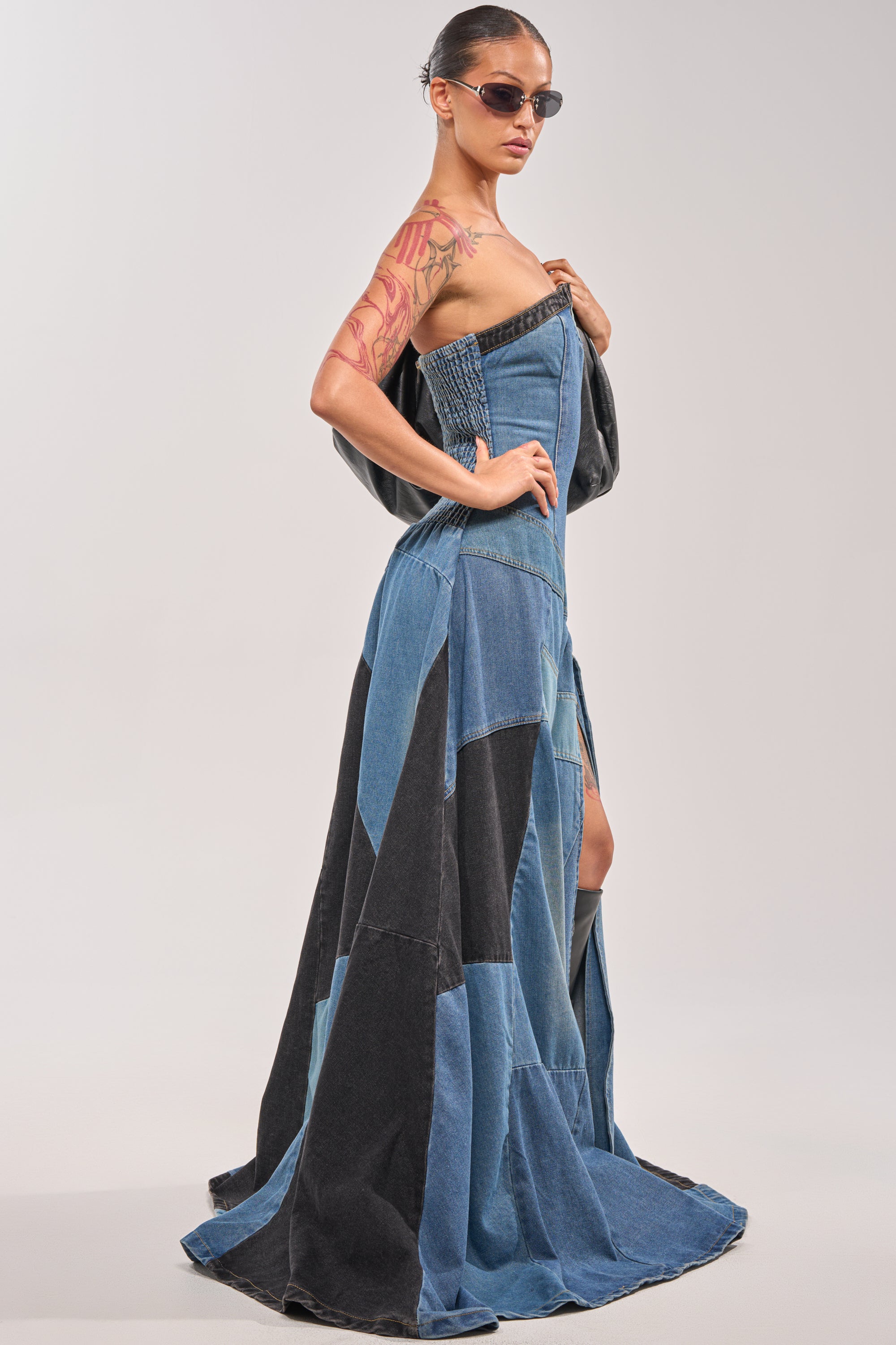 Wearing the LIFE IN DENIM STRAPLESS MAXI DRESS, someone poses against a plain light background with sunglasses and a colorful tattoo visible on their upper arm. 3