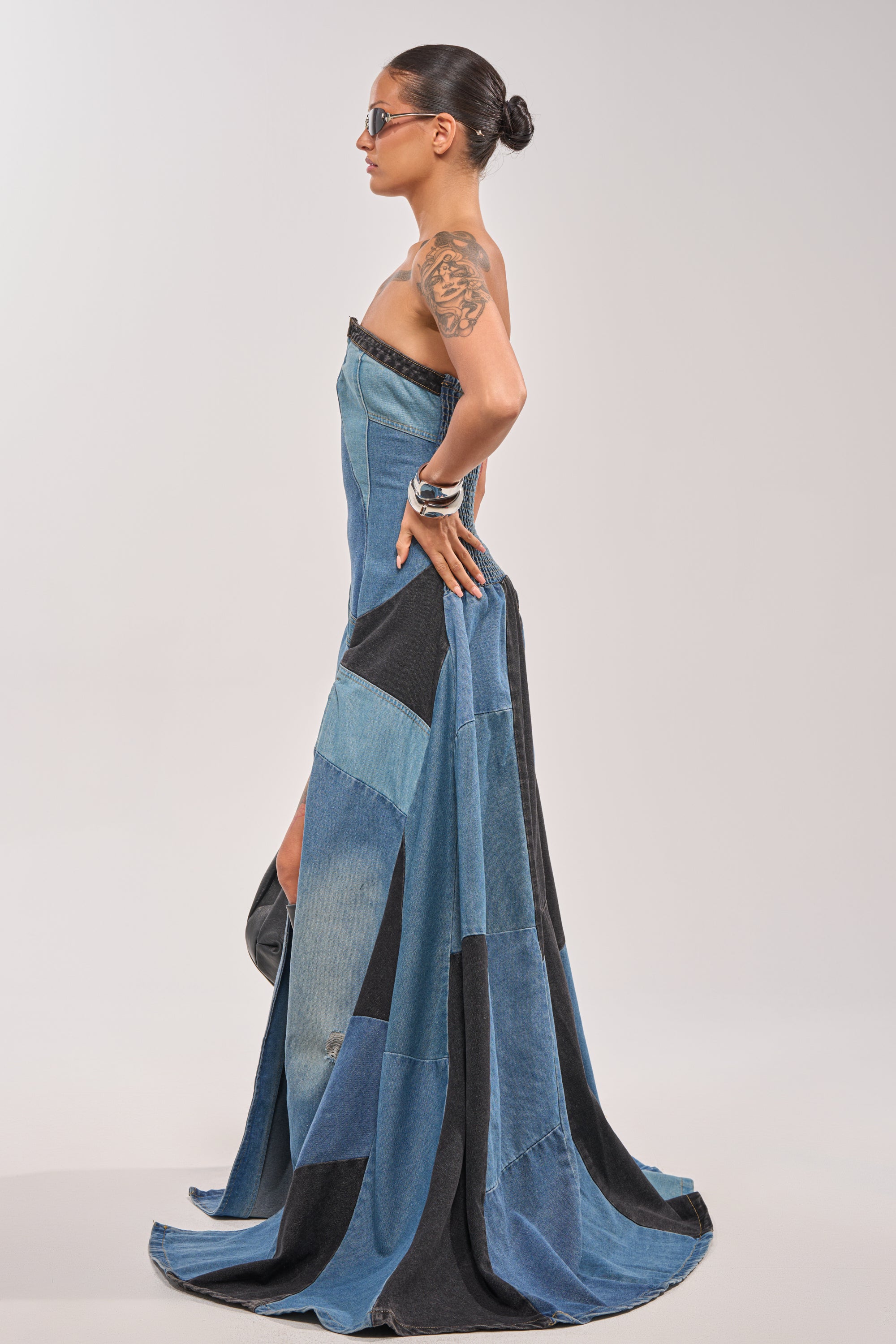 A woman in sunglasses stands in profile against a plain background, showcasing her upper arm tattoo while wearing the LIFE IN DENIM STRAPLESS MAXI DRESS and holding the back of her evening wear. 4