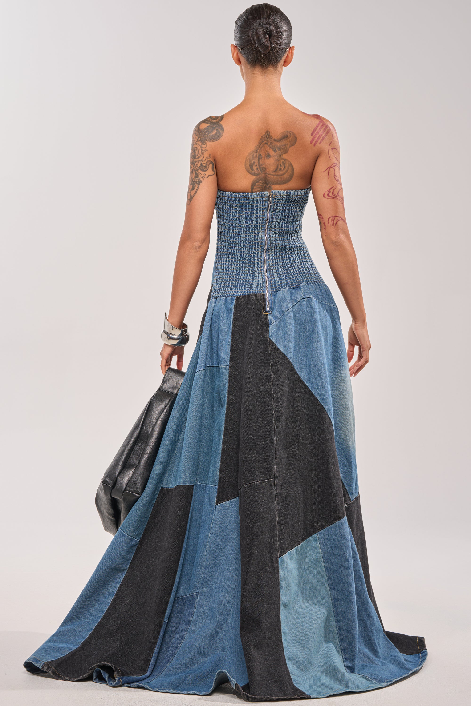 Wearing the LIFE IN DENIM STRAPLESS MAXI DRESS, a tattooed individual with jewelry is seen from behind, holding a black jacket in one hand. The fitted bodice and flared skirt make it ideal for standout evening wear. 5