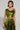 A person with short hair wears a shiny green blouse from the Metallic Dream collection, featuring a stylish cropped silhouette paired with a matching skirt. They have tattoos adorning their arms and torso, accessorize with a metallic clutch, hoop earrings, and a bracelet against a plain background.