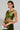 A woman in a green, sleeveless METALLIC DREAM BLOUSE and matching skirt poses against a plain background. She has short dark hair, numerous tattoos on her arms, and holds a shiny, abstract accessory in her hands.