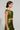 A person is shown in profile wearing the METALLIC DREAM BLOUSE, featuring a shiny green sleeveless top paired with a matching skirt. Their short dark hair complements the tattoos visible on their arm and midriff, highlighting the cropped silhouette. The background remains plain and neutral.