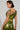 A person with short, dark hair is dressed in the METALLIC DREAM BLOUSE and coordinating metallic green trousers featuring a cropped silhouette. They have tattoos on their arm and are gazing to the side against a simple, light-colored background.