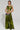 A person poses in a vibrant green two-piece ensemble, featuring the METALLIC DREAM BLOUSE with its distinctive asymmetrical design. They carry a black handbag accented with white details, and their arm is decorated with a tattoo, all set against a simple, light backdrop.