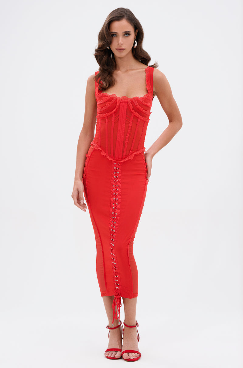 A woman with wavy brown hair models the LET ME REMIND YOU LACE UP MESH MIDI DRESS IN RED, featuring a corset-inspired bodice and lace-up detail, paired with matching red high heels against a plain white background.