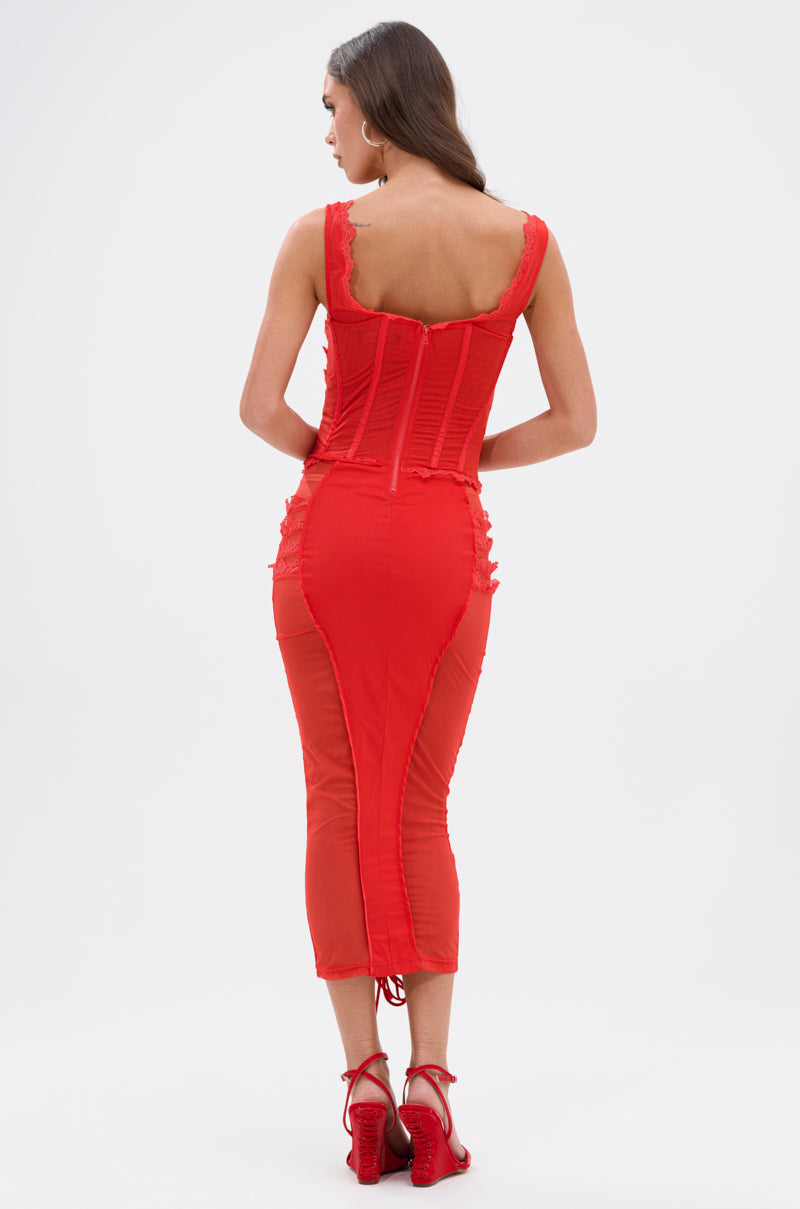 A woman with long brown hair faces away, wearing the LET ME REMIND YOU LACE UP MESH MIDI DRESS IN RED. The fitted, sleeveless dress has a corset-inspired bodice and lace design. She pairs it with red heeled sandals against a white background.