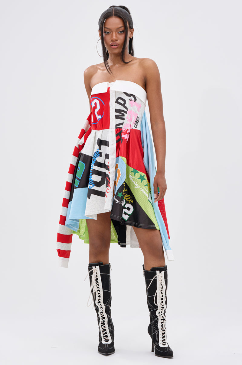 A woman faces forward in the STAND OUT GRAPHIC MINI DRESS, featuring a colorful patchwork of prints and stripes, paired with black knee-high lace-up boots with white laces. The background is plain white.