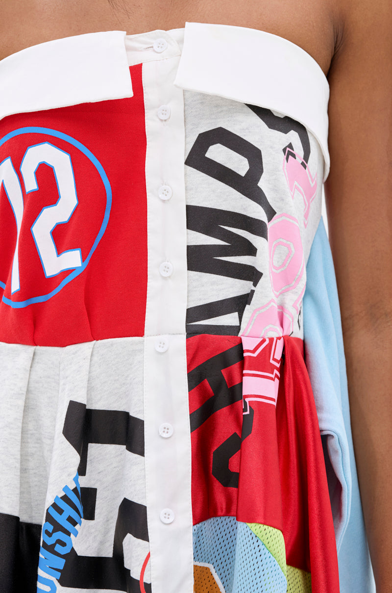 A close-up of the STAND OUT GRAPHIC MINI DRESS, a patchwork mini dress made from T-shirt fabric with printed graphics, numbers, colorful panels, and a white button-down front. The dress is worn off-shoulder.