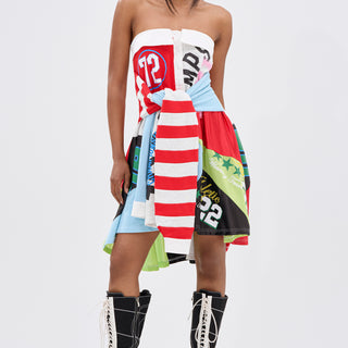 A woman stands confidently in the STAND OUT GRAPHIC MINI DRESS, featuring vibrant fabric patches with bold stripes and numbers, paired with black knee-high lace-up boots—a fresh street style look set against a plain white background.