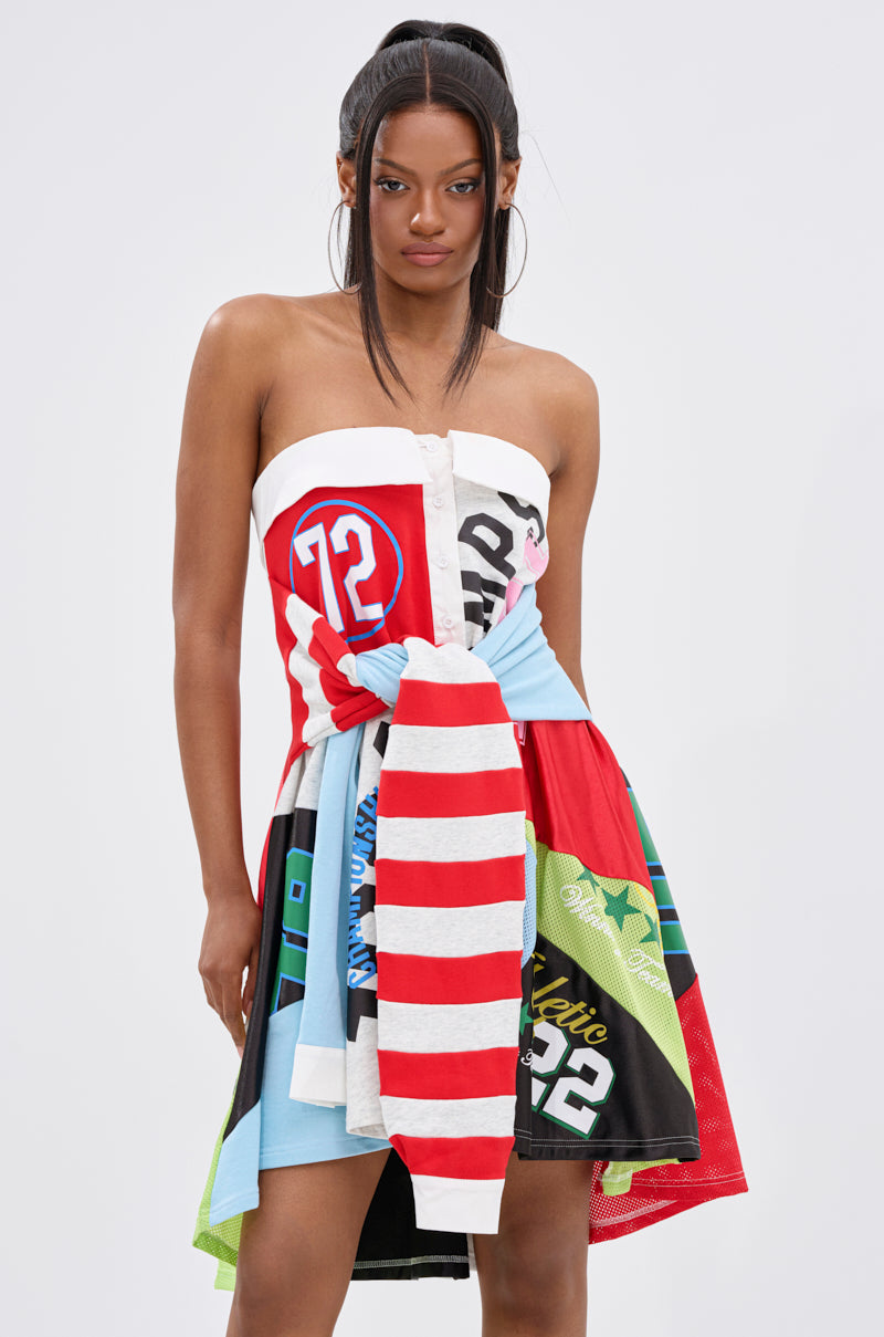 A woman with straight dark hair in a half-up ponytail wears the STAND OUT GRAPHIC MINI DRESS, a vibrant patchwork piece with numbers, stripes, and graphics, finished with a bold red-and-white striped bow at the waist.