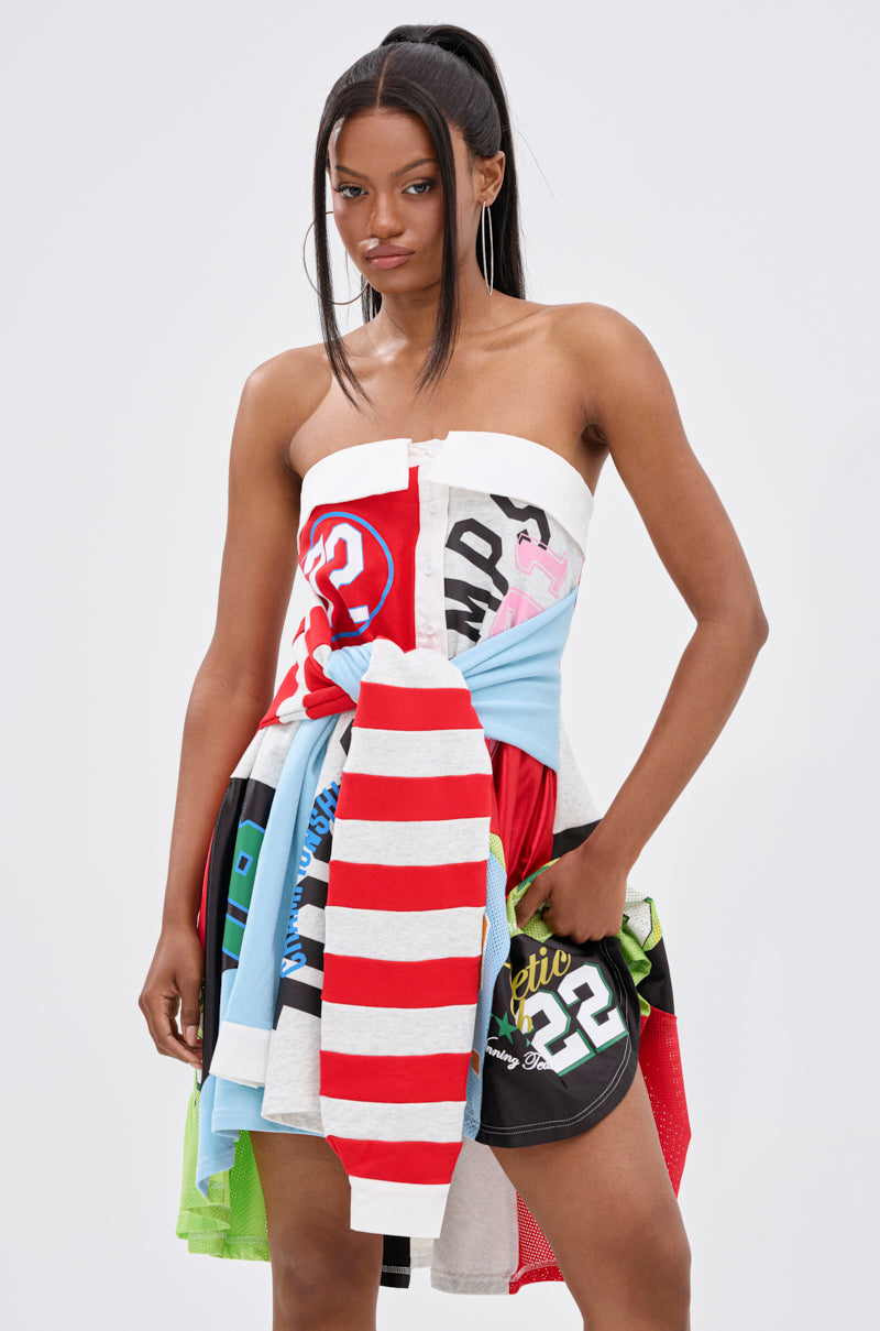 A woman with long straight hair in a high ponytail wears the STAND OUT GRAPHIC MINI DRESS, a strapless piece made from colorful, mixed-patterned fabrics featuring stripes, numbers, and graphic prints, standing confidently against a plain background.