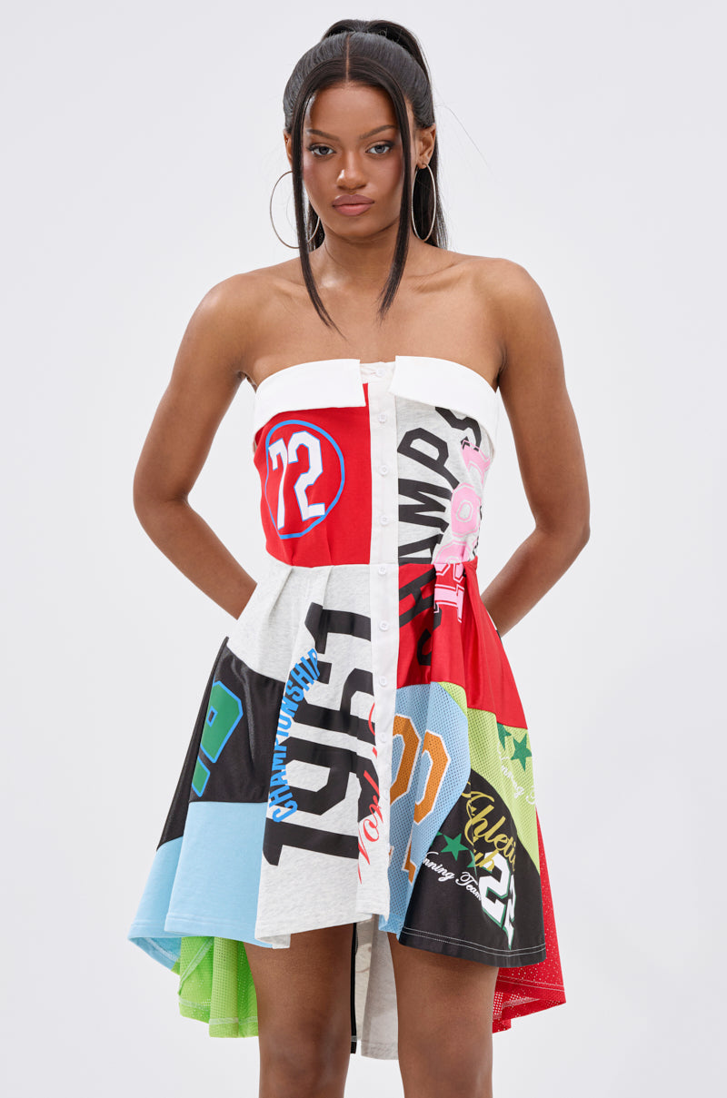 A woman poses hands on hips in the STAND OUT GRAPHIC MINI DRESS, a bold strapless mini crafted from colorful repurposed sports jerseys, set against a plain white background.