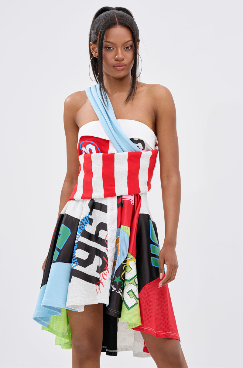A woman poses against a plain background in the STAND OUT GRAPHIC MINI DRESS, a bold one-shoulder mini with vibrant patterns and a handkerchief hem. Her long, straight hair is styled half-up for a trendy street style look.