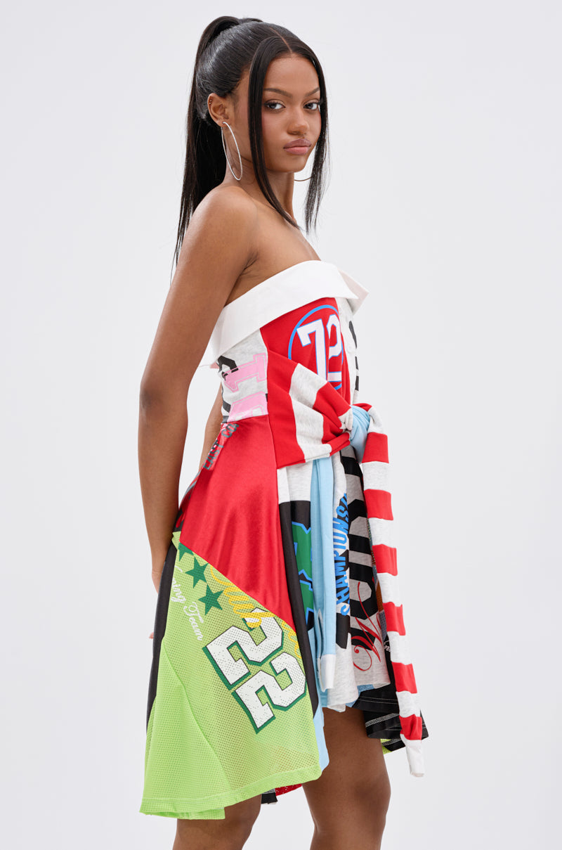 A woman with long straight hair wears the STAND OUT GRAPHIC MINI DRESS, a vibrant patchwork made from sports jerseys with numbers, stars, and bold patterns, standing against a plain light gray background.
