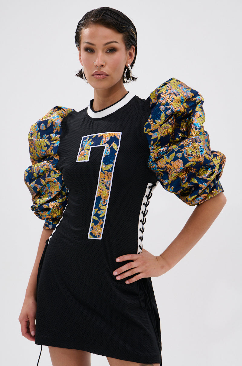 A woman stands confidently with one hand on her hip, wearing the VIBIN ON THE SIDELINES JERSEY MINI DRESS featuring a bold white number 7 and dramatic puffy Metallic Floral Brocade sleeves against a plain background. 0