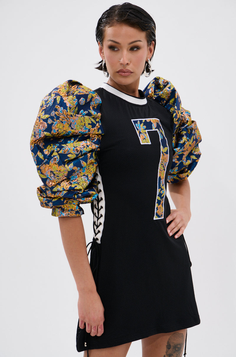 A woman stands with one hand on her hip in the VIBIN ON THE SIDELINES JERSEY MINI DRESS, featuring dramatic metallic floral brocade puffy sleeves and a bold number 7, striking a pose against a plain white background. 1