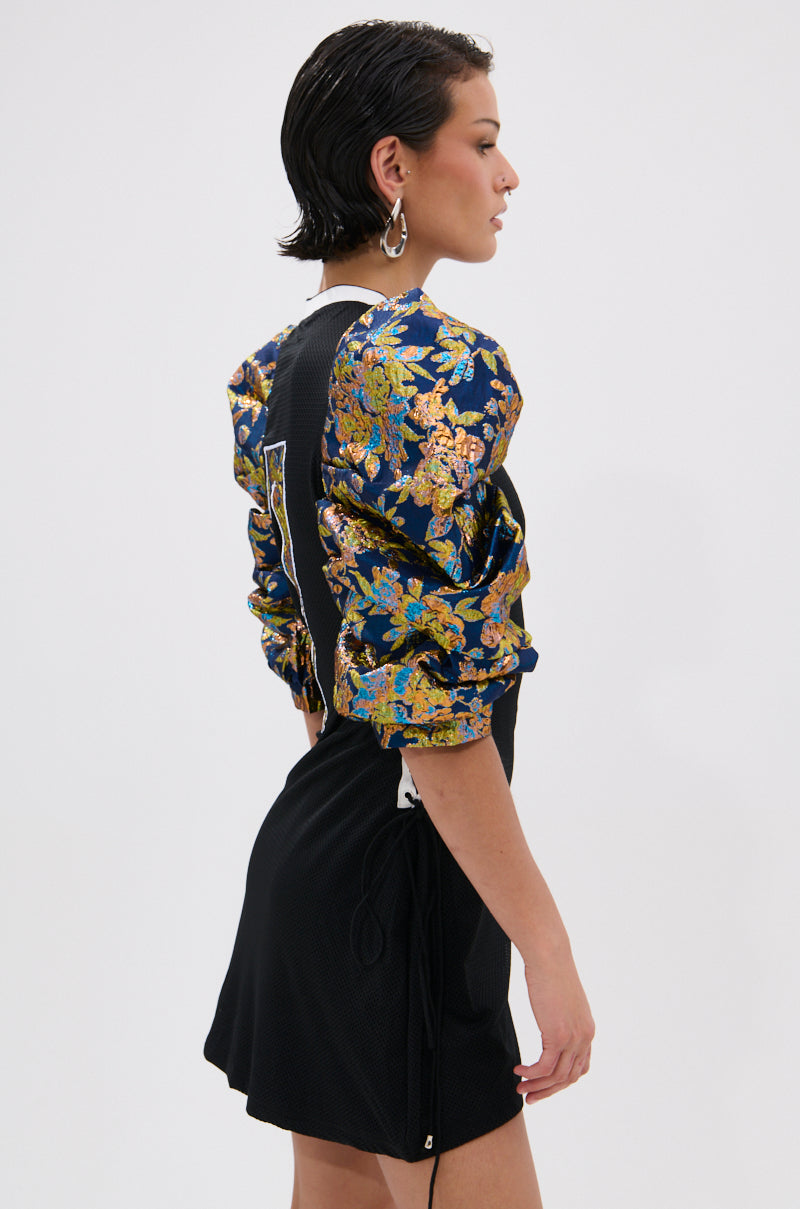 A woman with short, wet-look hair models the VIBIN ON THE SIDELINES JERSEY MINI DRESS, featuring puffed blue and gold metallic floral brocade sleeves. She stands sideways to highlight the dress’s textured fabric and ornate sleeve detail. 2