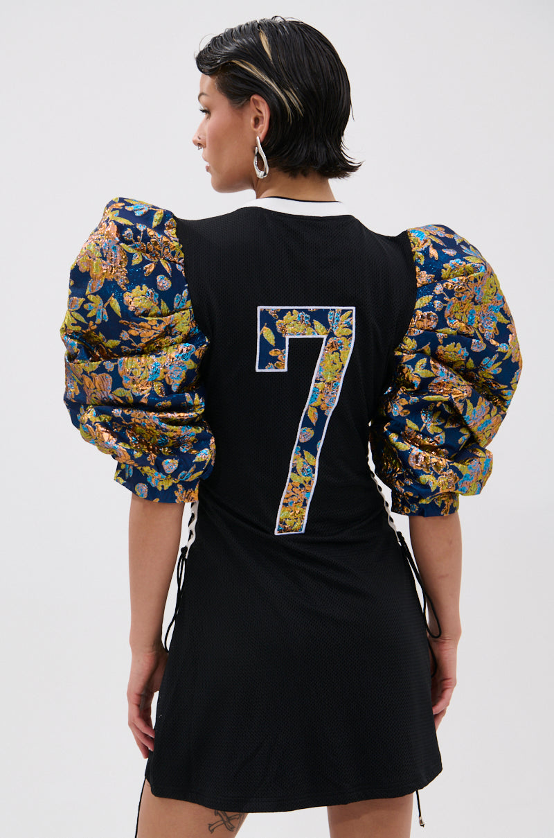 A person faces away, Vibin’ On The Sidelines in the black VIBIN ON THE SIDELINES JERSEY MINI DRESS featuring puffy metallic floral brocade sleeves and a floral-print number 7 on the back. Short dark hair and large hoop earrings complete the look. 3