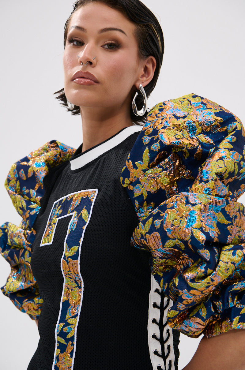 A woman with short dark hair and hoop earrings wears the VIBIN ON THE SIDELINES JERSEY MINI DRESS, which features a large “T” on the front and dramatic, metallic floral brocade puff sleeves. She gazes confidently at the camera. 5