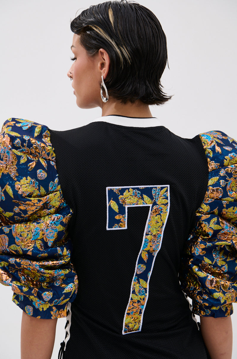 A person with short dark hair and silver earrings is seen from behind, wearing the VIBIN ON THE SIDELINES JERSEY MINI DRESS featuring floral puffed sleeves in metallic brocade and a bold patterned number 7 on the back. 6