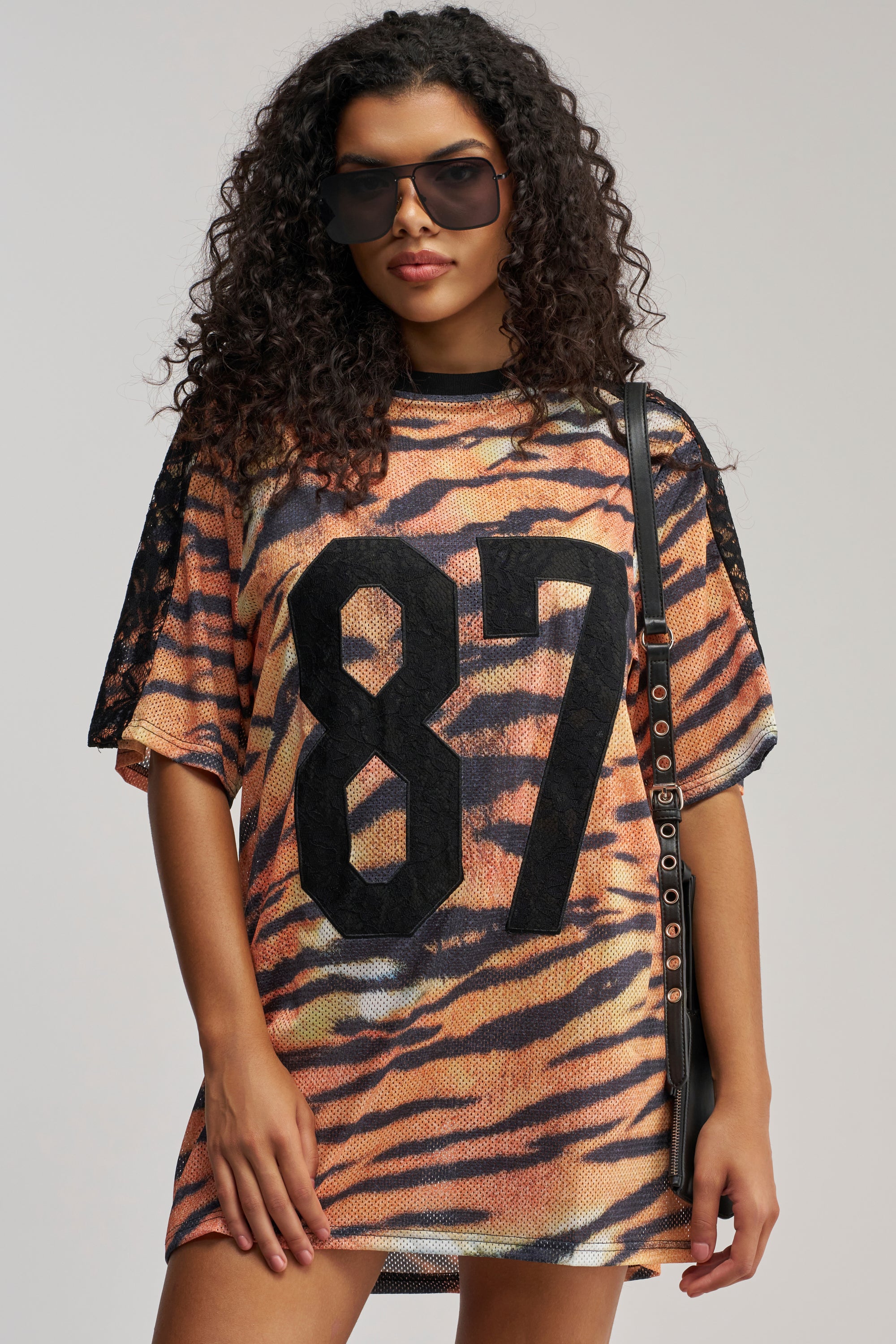 A person stands against a plain background wearing the SHE FEISTY LACE JERSEY MINI DRESS—a vibrant orange tiger print mini dress with the number 87 and black lace detailing—paired with sunglasses and a black strap bag.