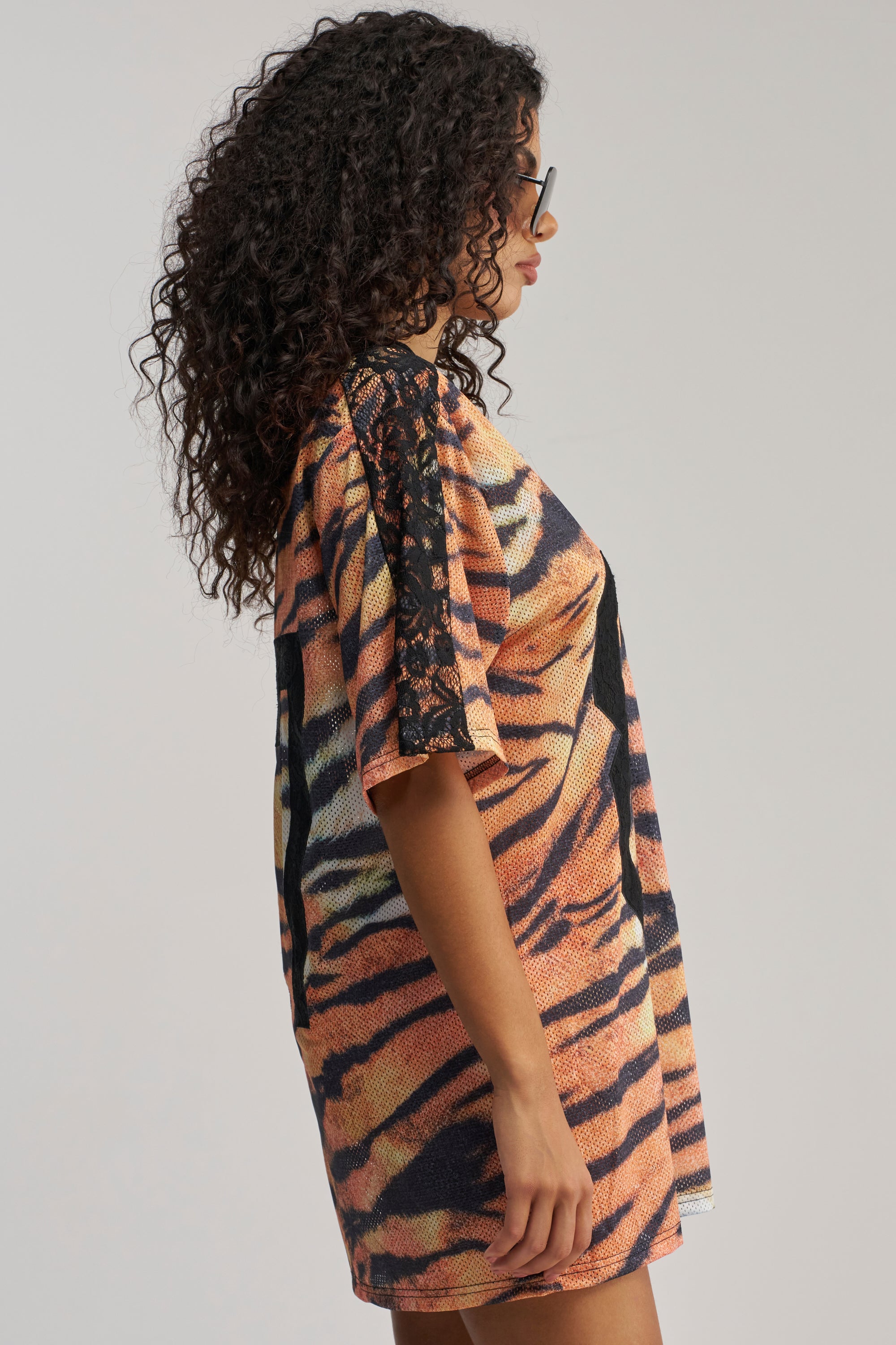A woman in sunglasses stands in profile against a plain background, wearing the SHE FEISTY LACE JERSEY MINI DRESS—a short orange tiger print dress featuring black lace detailing on the sleeves.