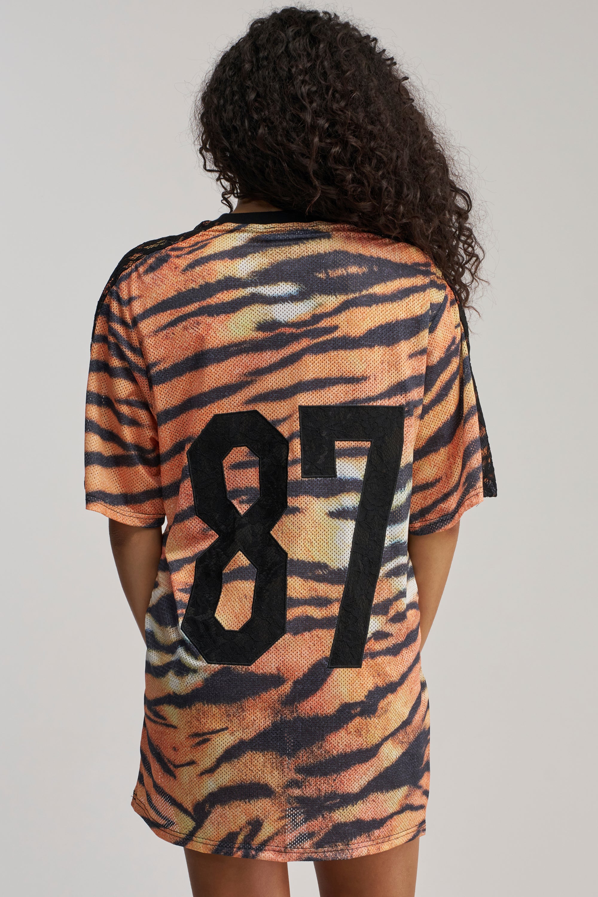 Wearing the SHE FEISTY LACE JERSEY MINI DRESS, a person with long curly hair faces away, showcasing the loose streetwear silhouette. The dress features an eye-catching orange and black tiger stripe pattern with the number 87 boldly printed in large black numerals on the back, while subtle black lace detailing adds a unique touch to its bold design.