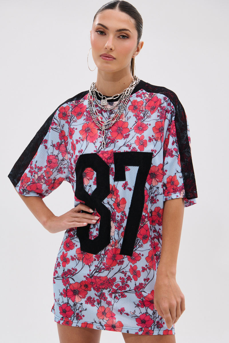 Wearing the POPPIN POPPIES LACE JERSEY DRESS with a bold poppy print and black "87" on the front, she pairs layered silver necklaces and hoops for an effortlessly sporty floral look against a plain background. 2