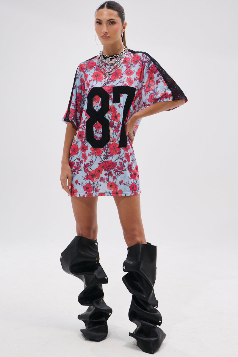 A woman stands confidently in the POPPIN POPPIES LACE JERSEY DRESS, featuring bold black "87" print. She completes the look with oversized slouchy black boots, slicked-back hair, and layered silver necklaces. 11
